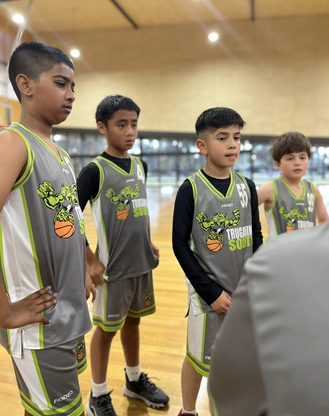 Young boys in basketball uniforms listening during a team meeting on an indoor basketball court.