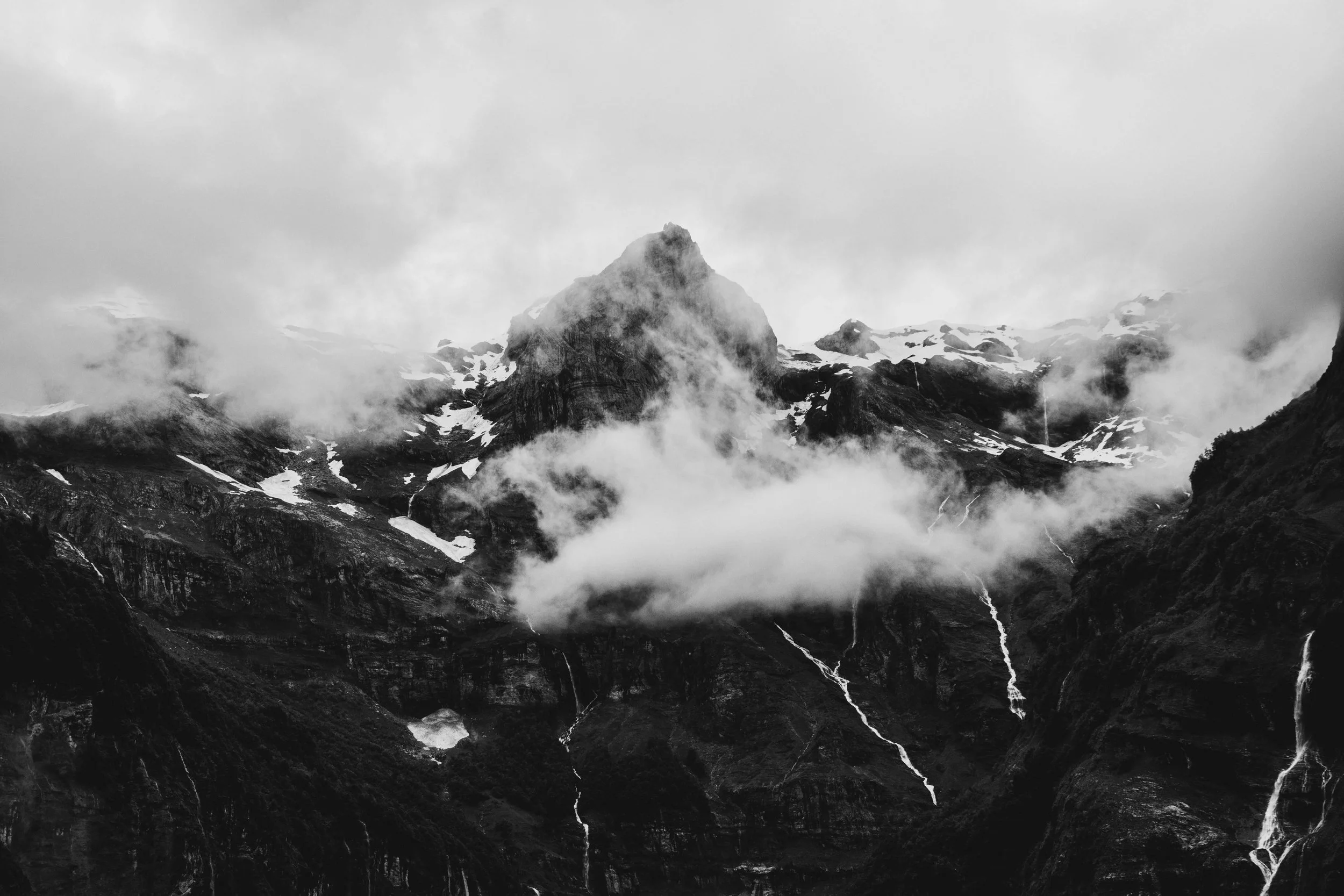 A black and white photograph of a mountain with snow patches on its slopes, surrounded by clouds and mist, with multiple waterfalls flowing down the dark rocky terrain.