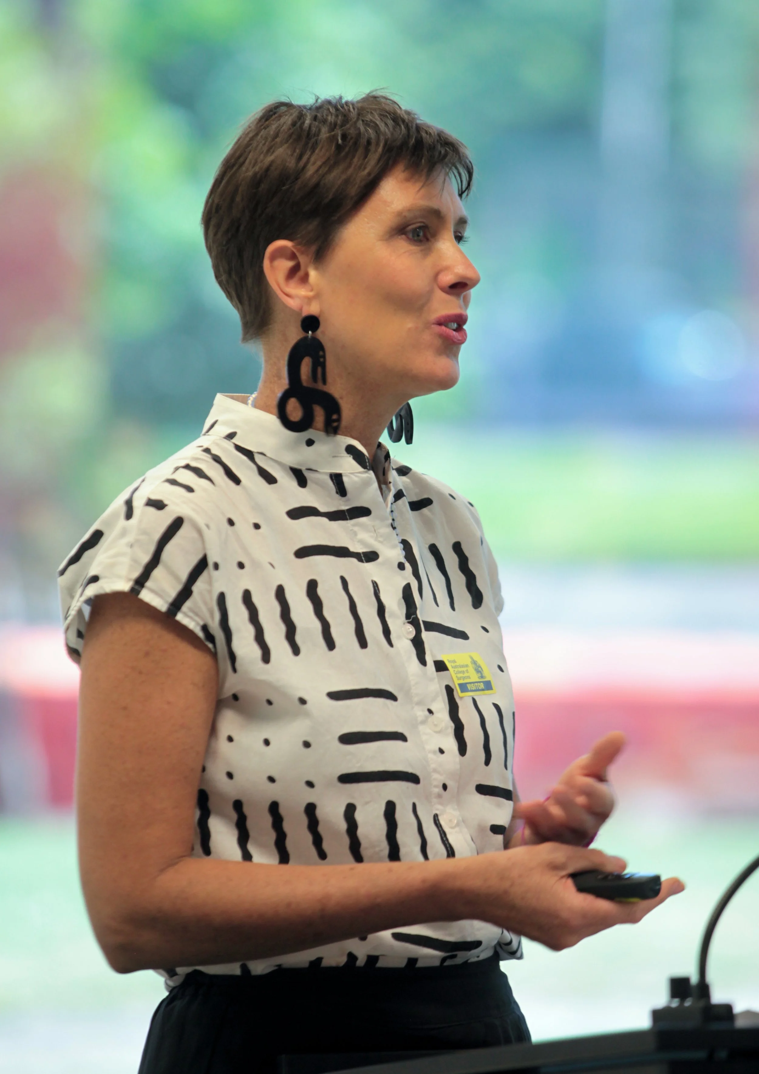 Woman with short brown hair wearing black and white patterned shirt and large black earrings, standing indoors with a colorful blurry background.