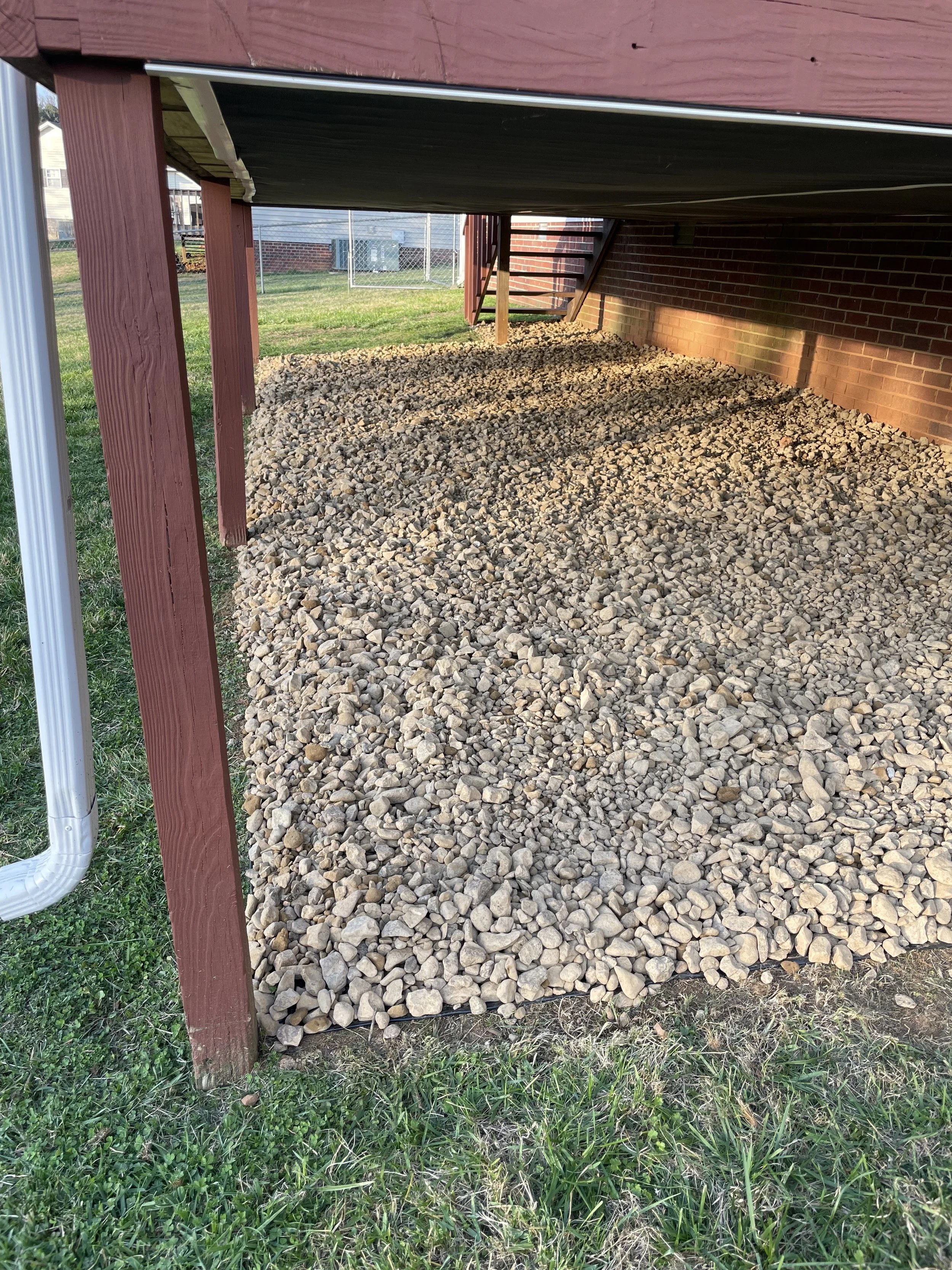 Area under a deck filled with small rocks, with stairs and brick wall visible in the background.