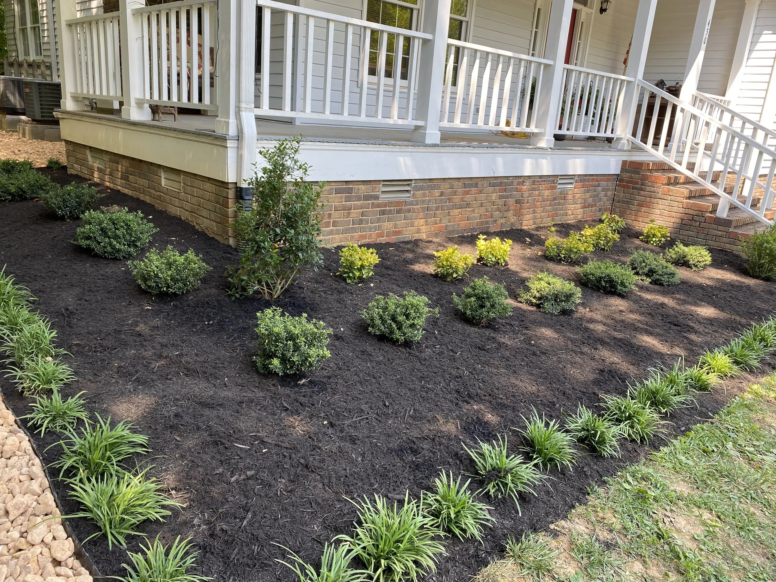 Front yard garden with neatly planted shrubs and flowering plants, mulch covering the soil, brick house foundation, white porch railing, and stairs.