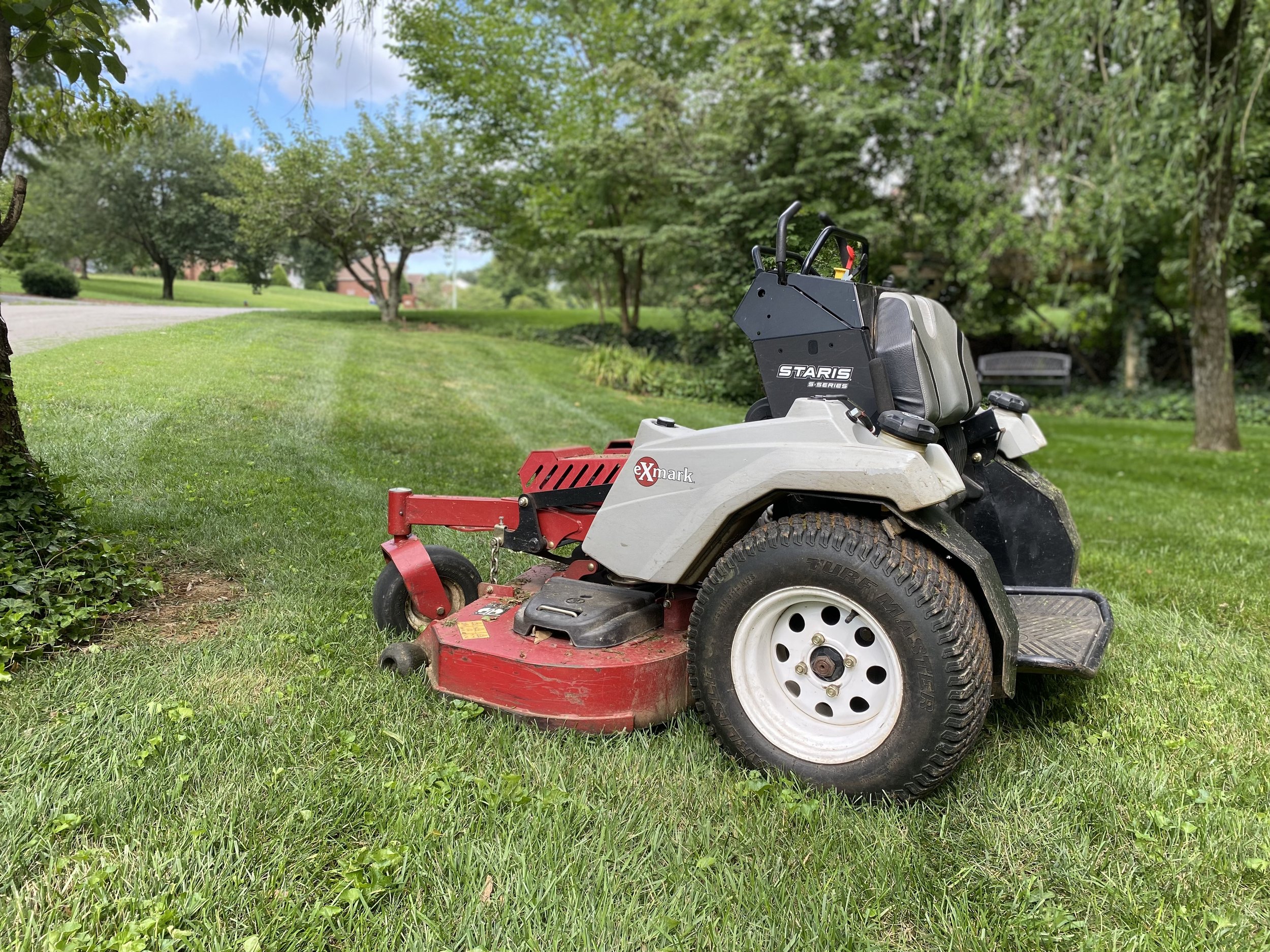 A ride-on lawn mower on a grassy lawn with trees and a bench in the background.
