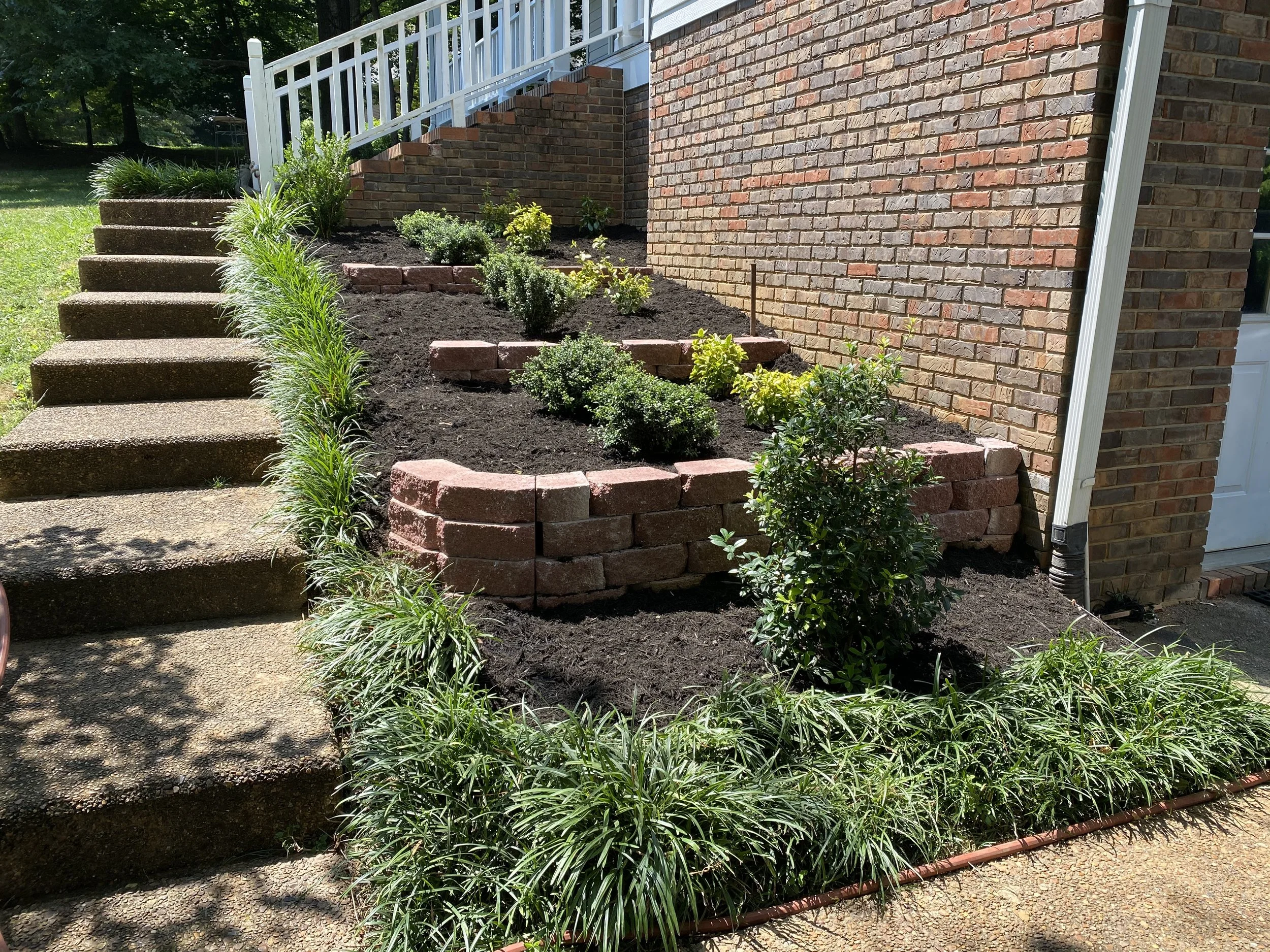 Raised garden bed with brick border next to brick house wall, filled with plants and soil, with concrete steps nearby.