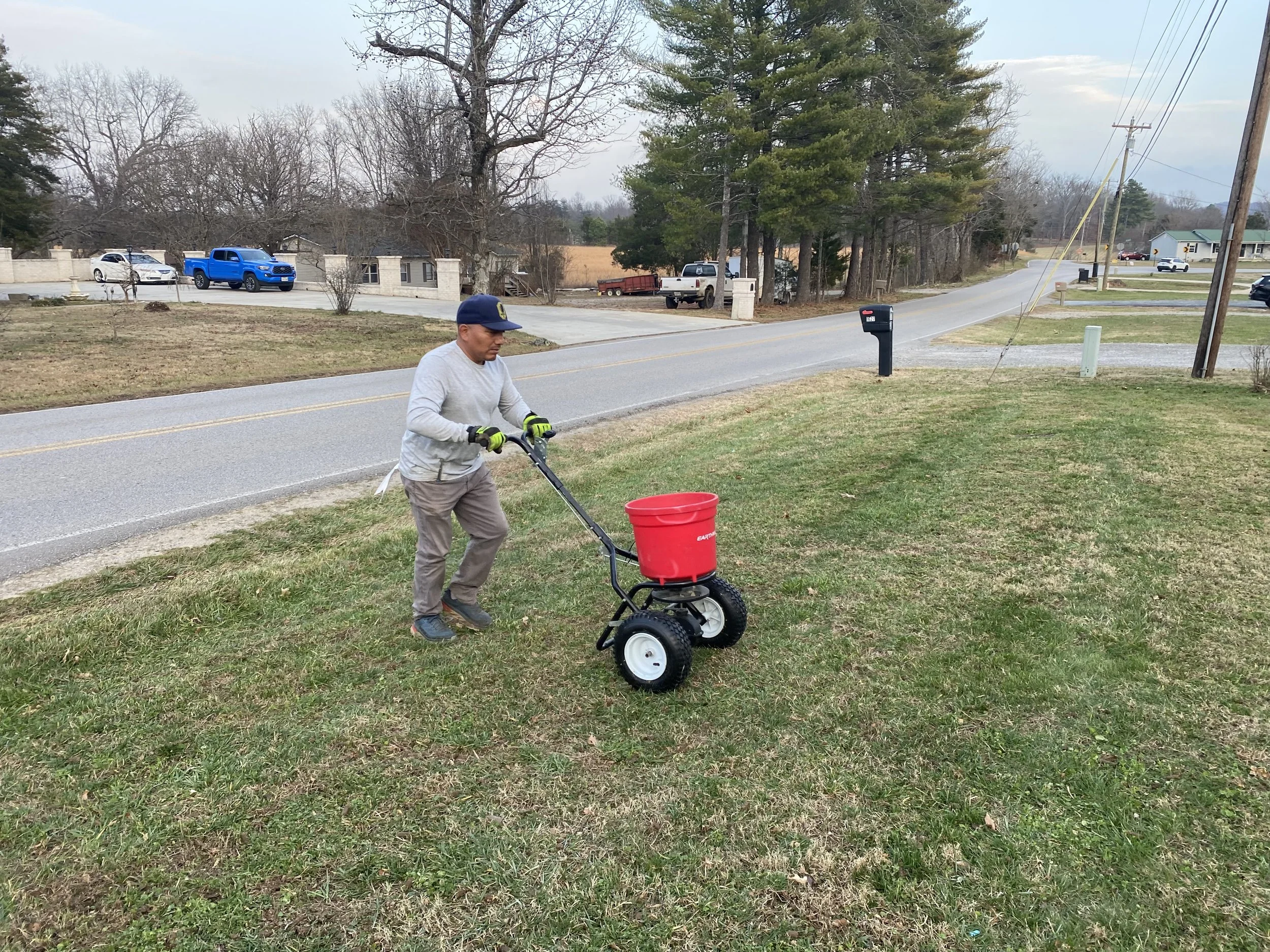 A man using a small motorized cart for blowing or sweeping leaves on a grassy area beside a street, with trees and parked cars in the background.