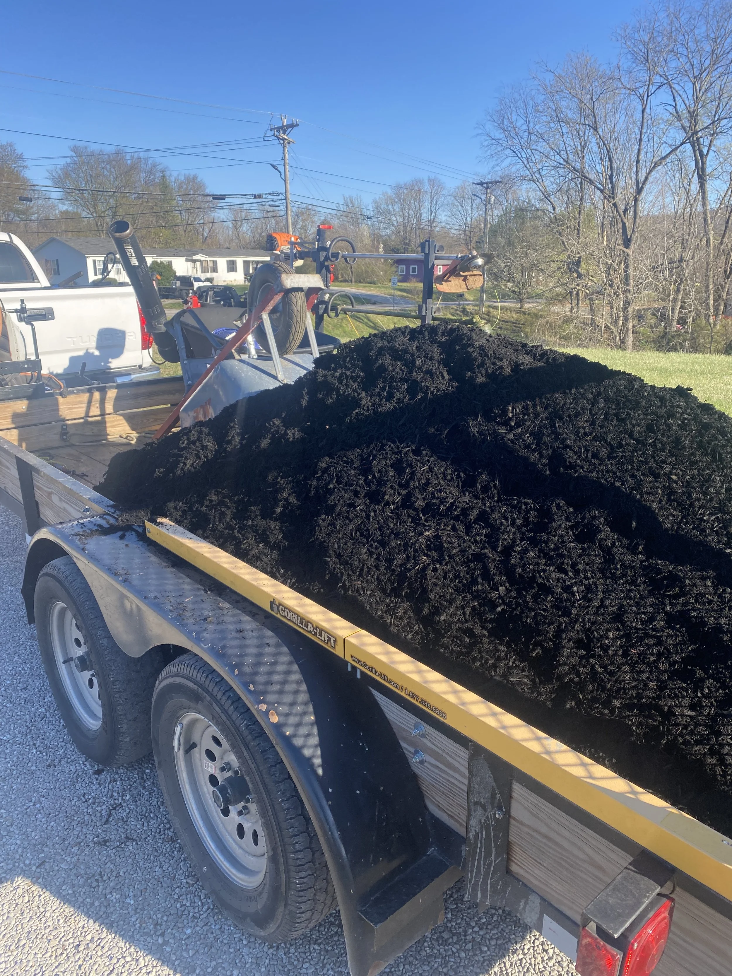 A trailer loaded with dark soil or compost, attached to a parked truck, outdoors on a clear day with trees and houses in the background.