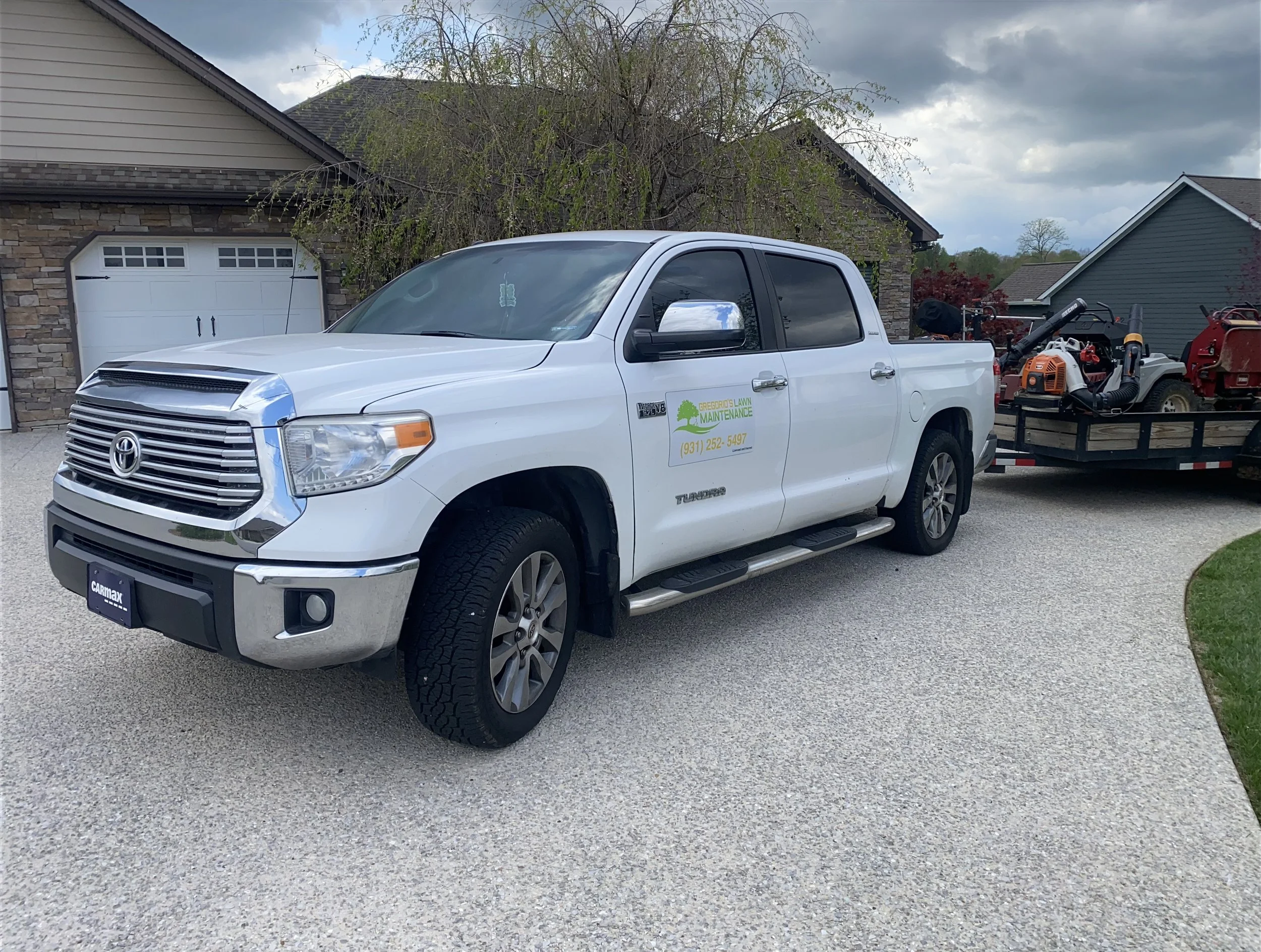 White Toyota Tundra pickup truck with green lawn maintenance company branding on the door, parked on a residential driveway. The truck is attached to a trailer carrying landscaping equipment, such as leaf blowers and other gardening tools.