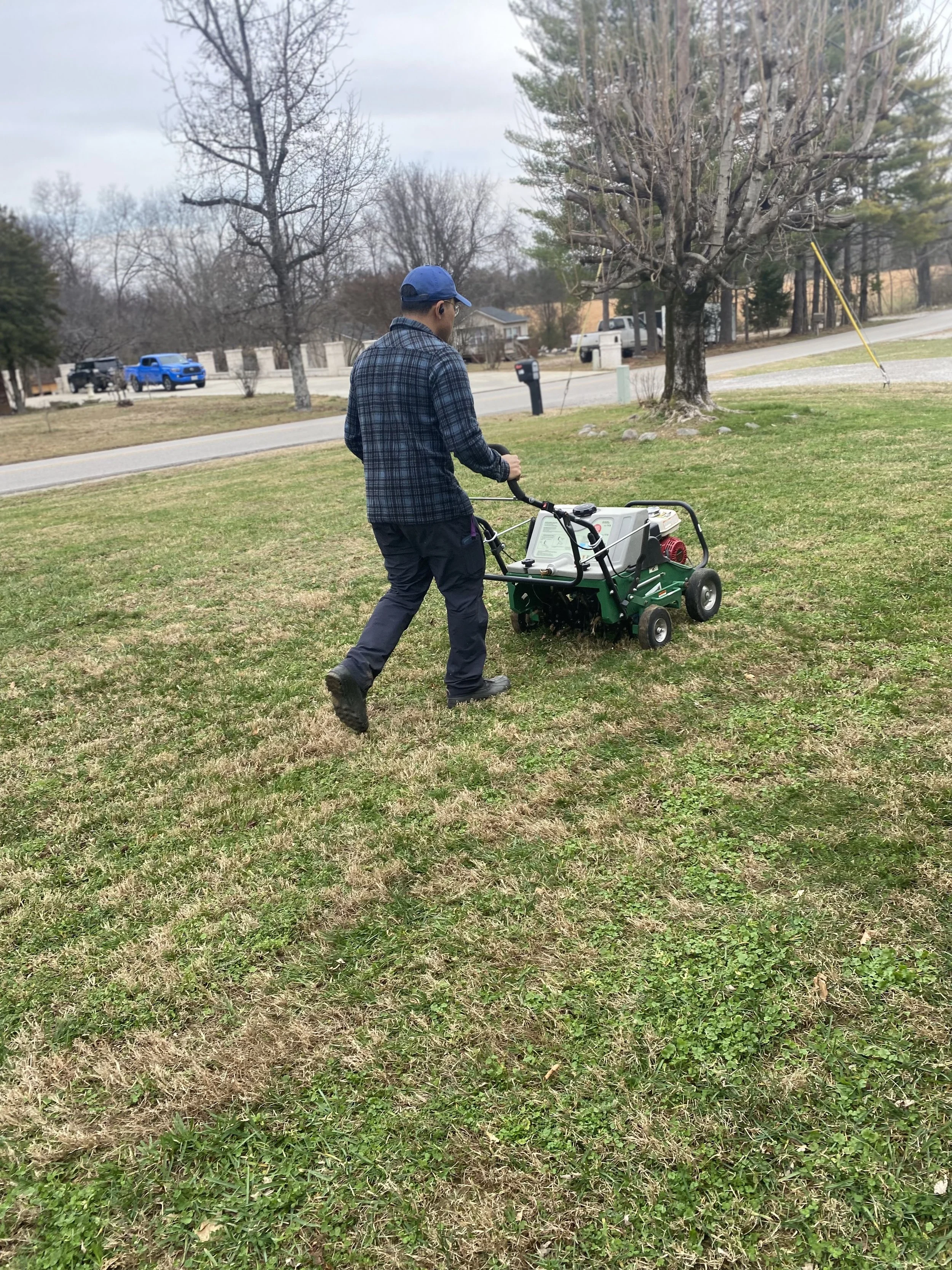A man in a blue cap and plaid jacket pushes a portable lawn aerator on a grassy yard.