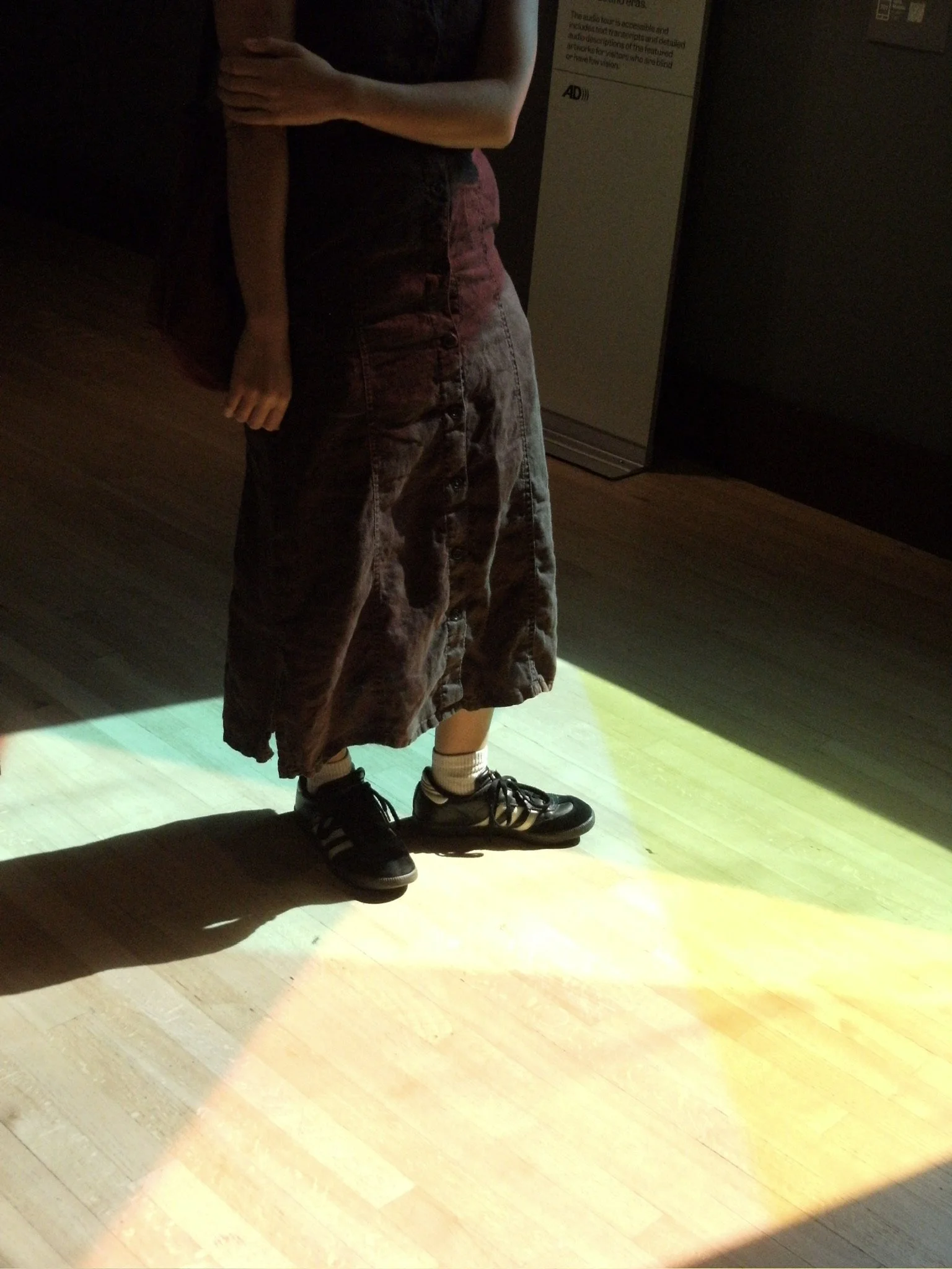 Reflection of a person in a mirror, showing their torso and legs, wearing a skirt and sneakers, standing on a wooden floor with rainbow beams of light.