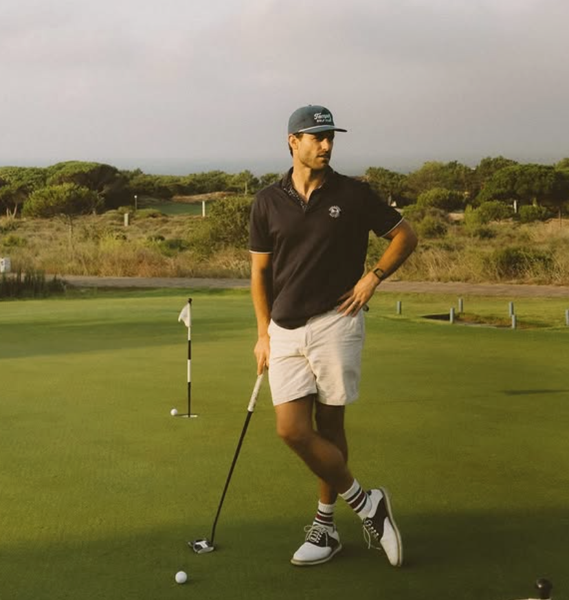 A man stands on a golf course putting on the green, holding a golf club, with a golf ball nearby, surrounded by trees and a cloudy sky.