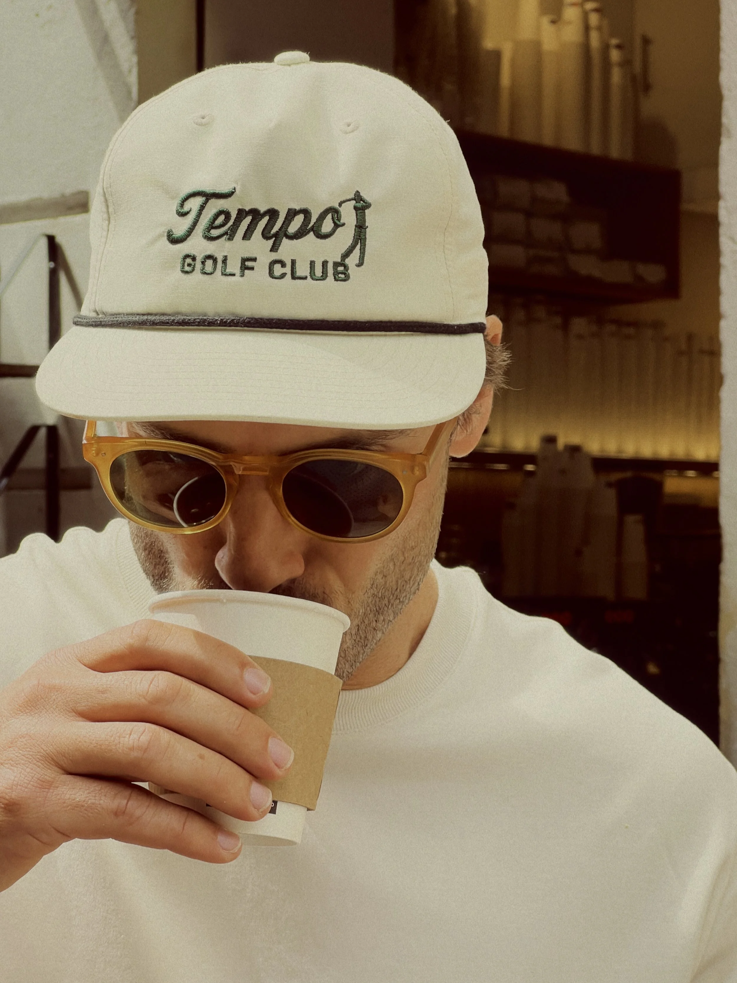A man wearing a white cap with 'Tempo Golf Club' embroidered on it, yellow-tinted sunglasses, and a white shirt, drinking from a paper cup inside a cafe.