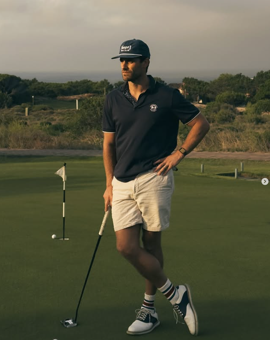 A man stands on a golf course putting green, holding a golf club, with a golf ball near a hole flag behind him, during sunset.
