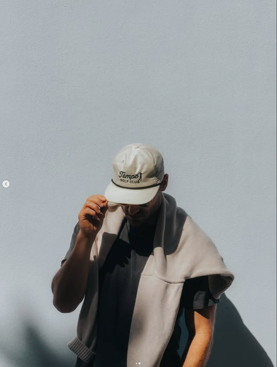 A person wearing a white cap and a beige hoodie with dark t-shirt underneath, standing against a plain light blue background, adjusting the cap.