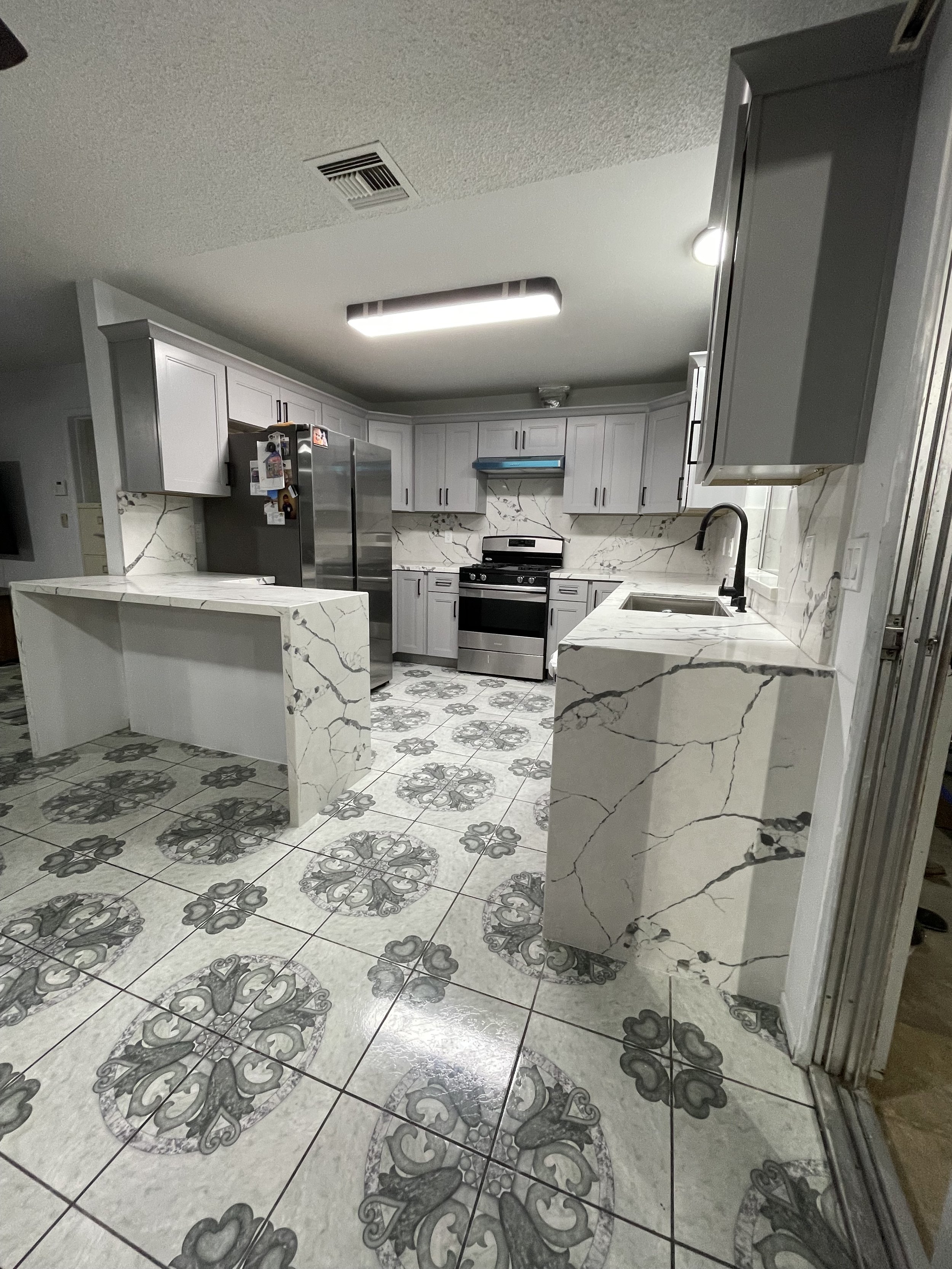 Modern kitchen with white marble countertops, white cabinets, and a patterned tiled floor. Contains a stainless steel refrigerator, stove, and sink.