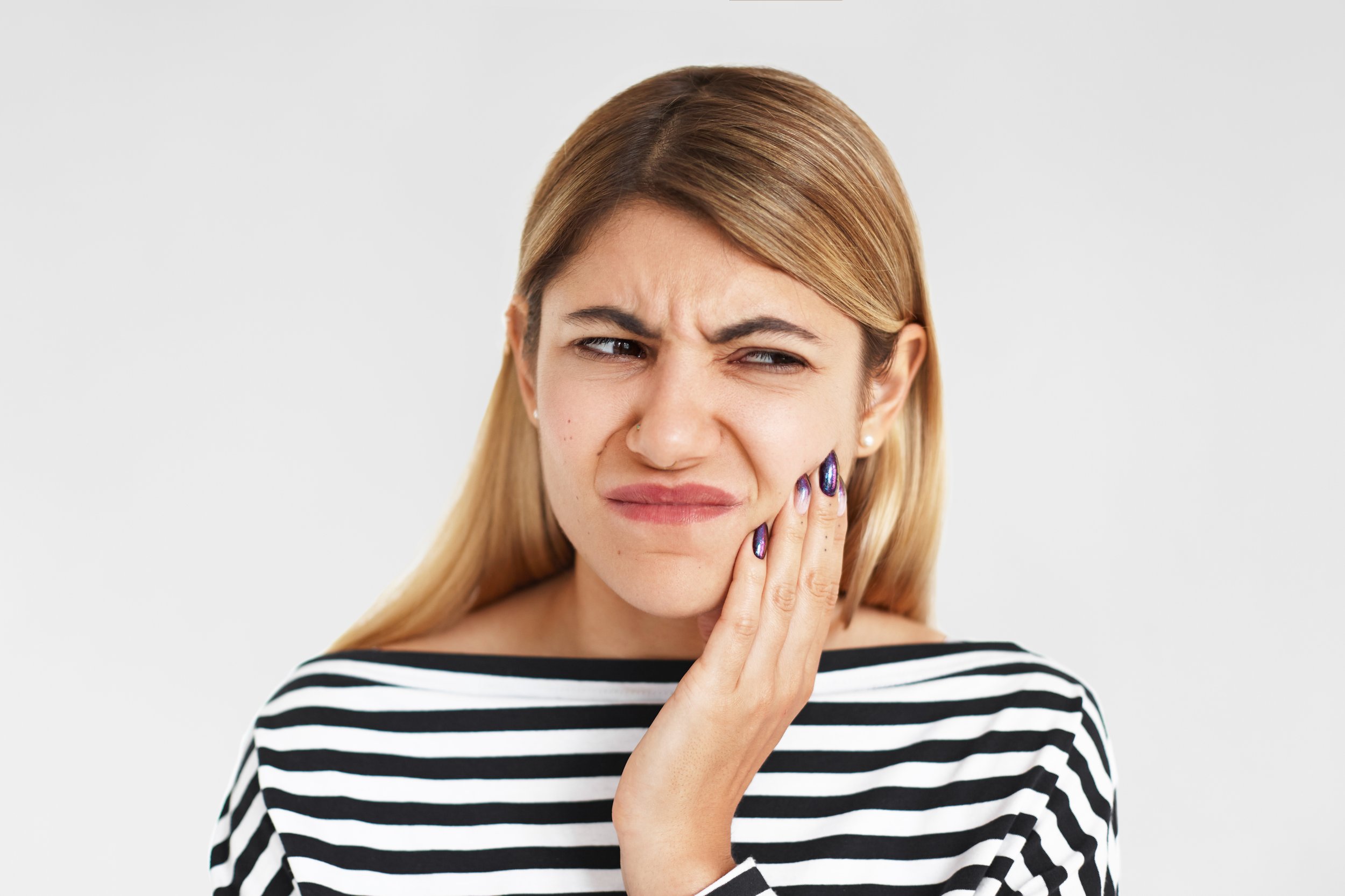 A woman with blonde hair, wearing a black and white striped shirt, touching her jaw with a pained or uncomfortable facial expression.