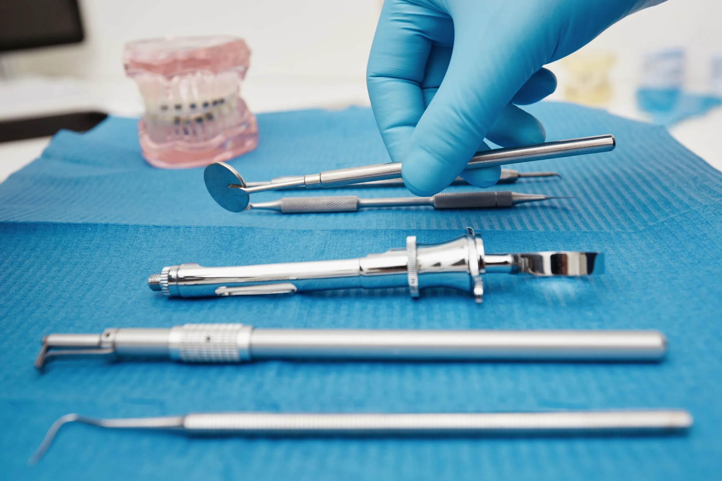 Dental tools arranged on a blue mat, including a dental mirror, probes, and other instruments, with a dental model in the background.
