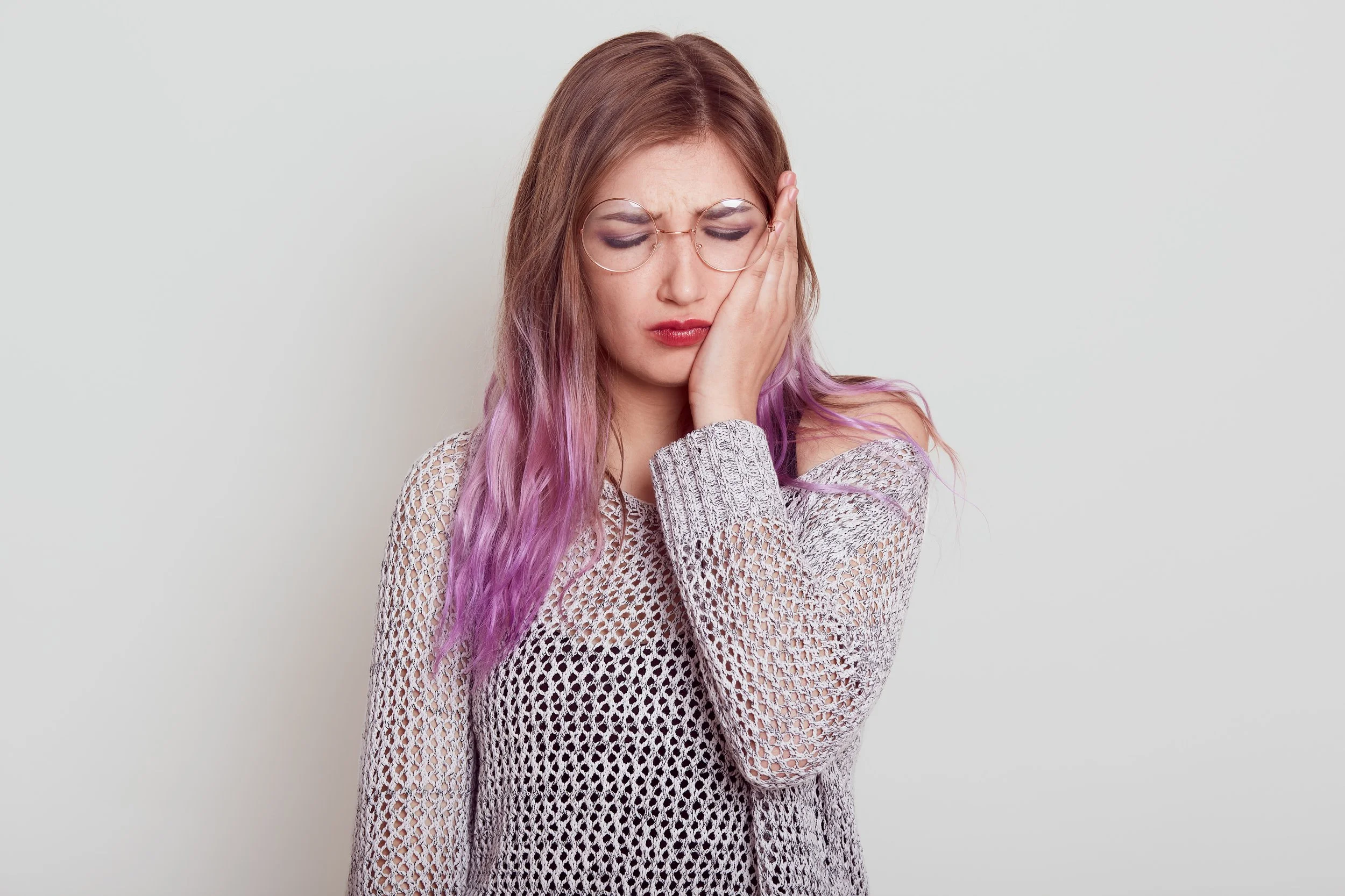 A young woman with long pink hair wearing glasses and a white knitted sweater, holding her face with a look of pain or frustration against a plain light background.