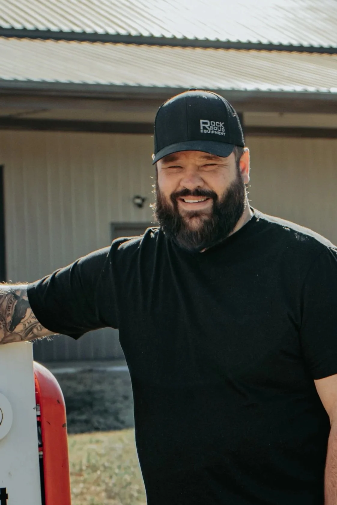 A smiling man with a beard wearing a black cap and black t-shirt, standing outdoors next to a fire extinguisher.