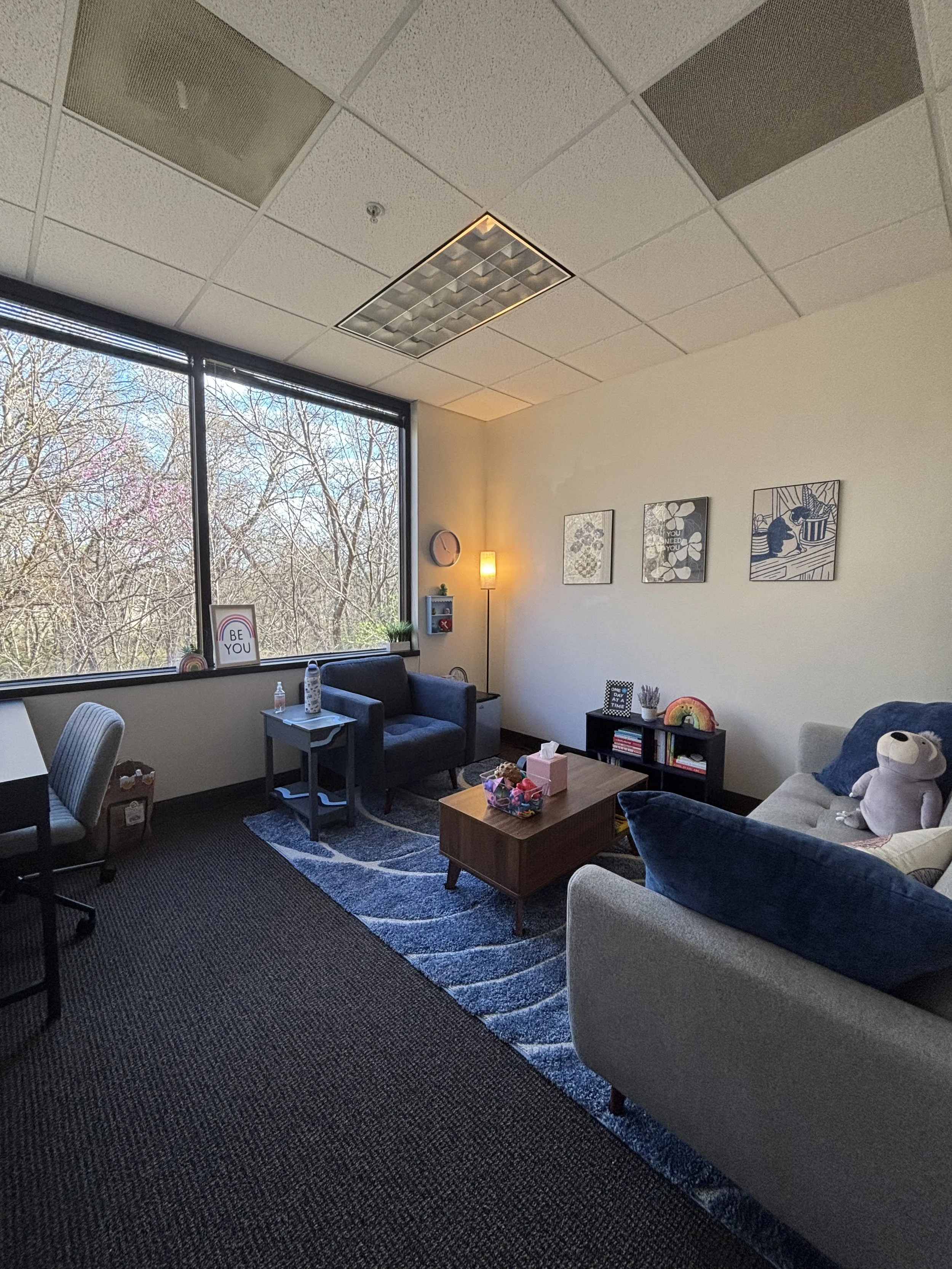 An office or therapy room with large window showing trees outside, a blue armchair, a beige sofa with colorful pillows and a stuffed animal, a coffee table with tissue box and toys, a small bookcase, wall art, a floor lamp, and a desk with a chair.