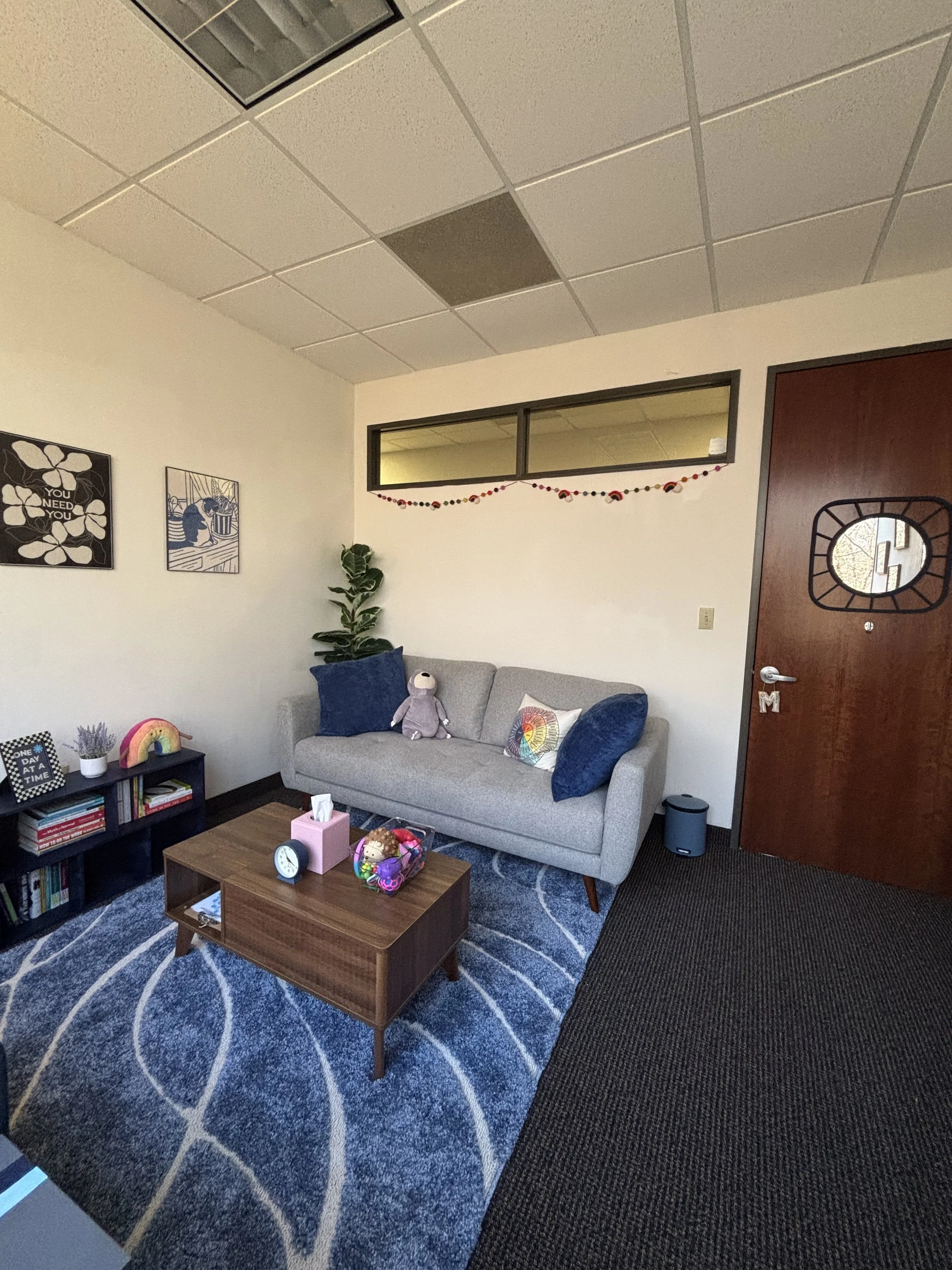A cozy office or lounge corner with a gray couch, colorful pillows, plush toys, a small bookshelf, and a wooden coffee table on a blue patterned rug. There are wall art, a potted plant, and a decorated door.