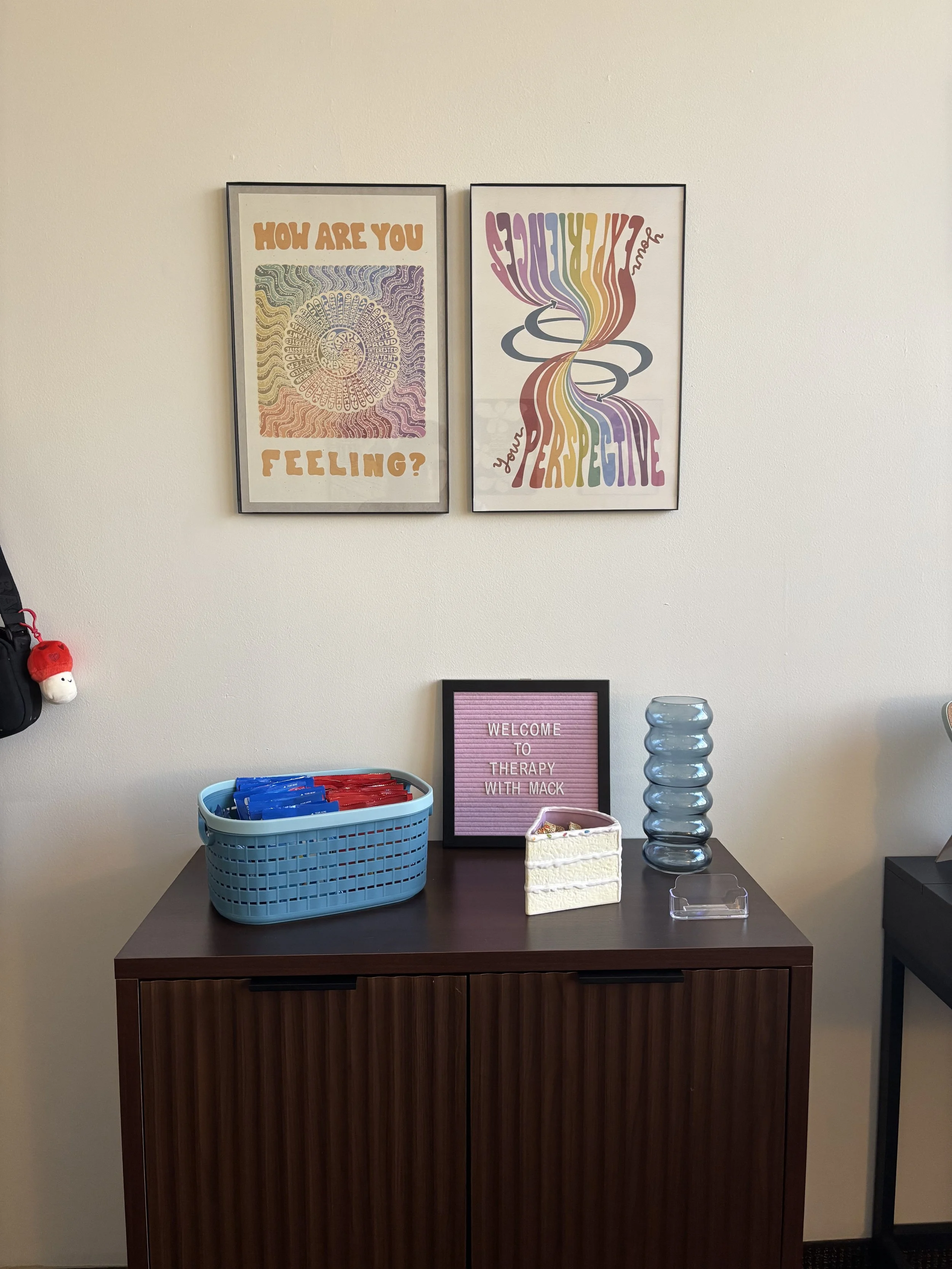 A therapy room corner with decorative posters on the wall, a dark brown cabinet with therapy supplies, a pink letter board reading 'Welcome to therapy with Mack,' a small white tissue box, a stack of clear blue and gray glass vases, and a small clear plastic container.