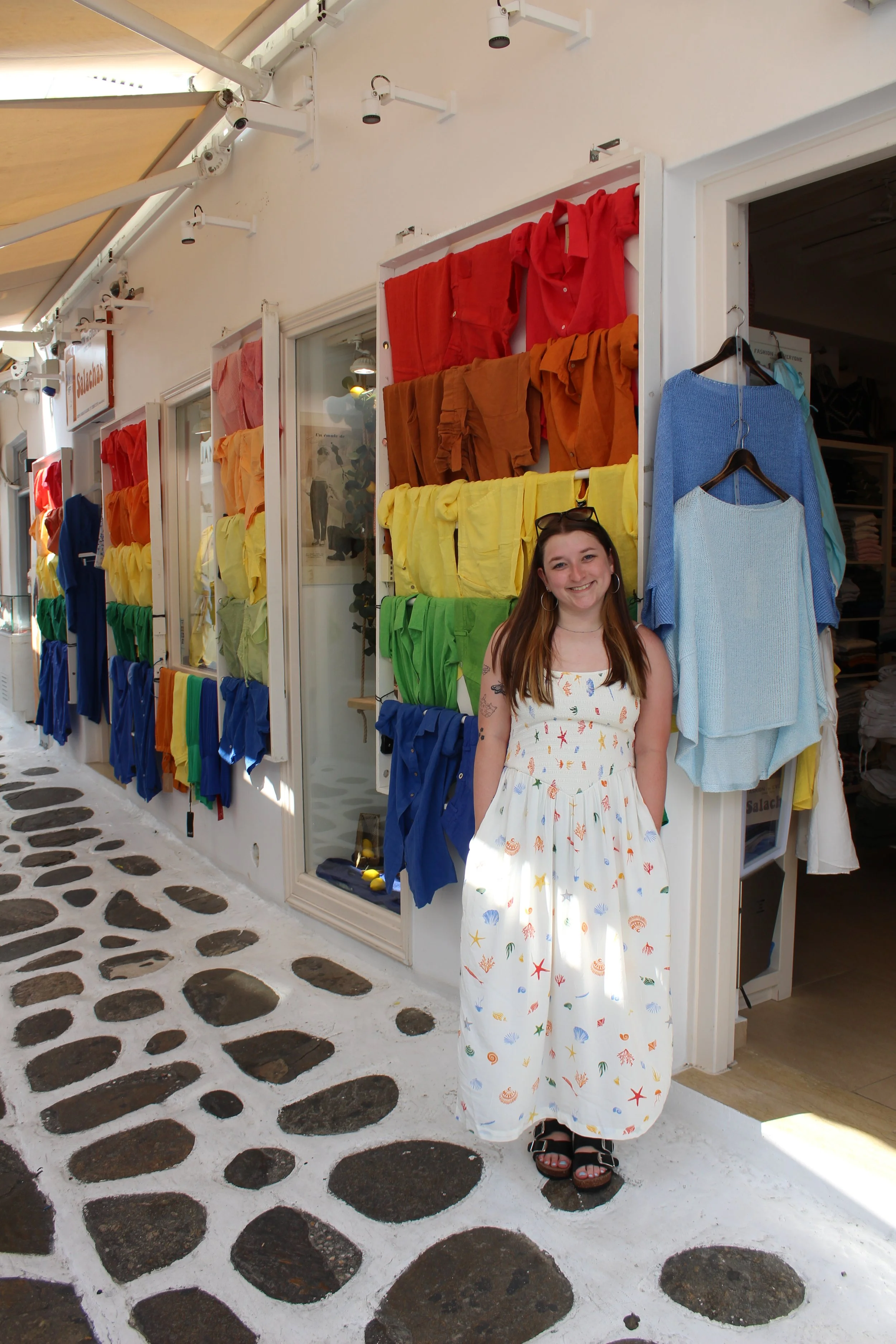 queer woman in a dress posing in front of a store with t shirts arranged in rainbow fashion, similar to a lgbtq+ pride flag