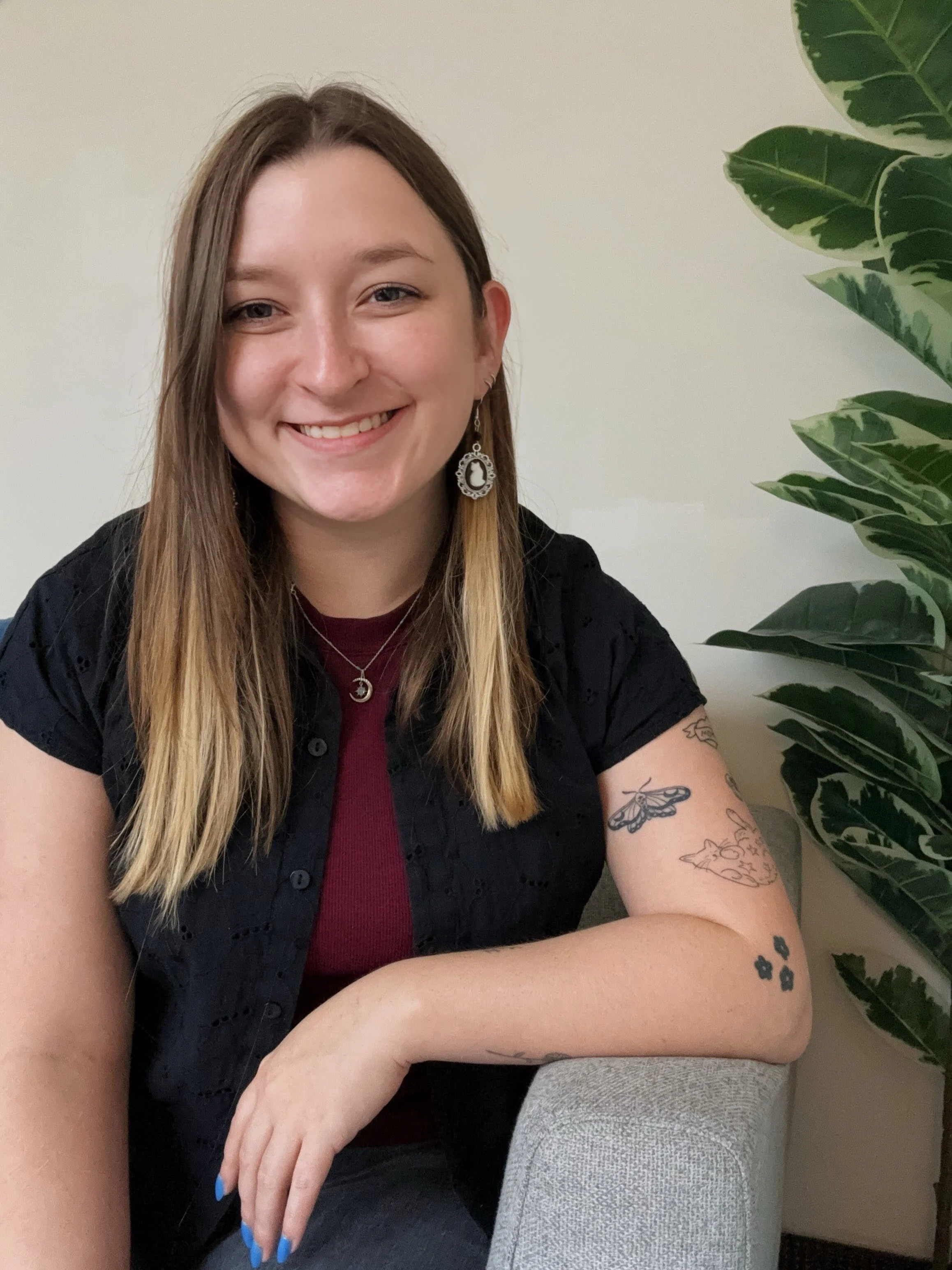 A smiling woman with shoulder-length light brown hair, wearing a black top, maroon shirt, and jewelry, sitting on a gray sofa with a large green leafy plant in the background.