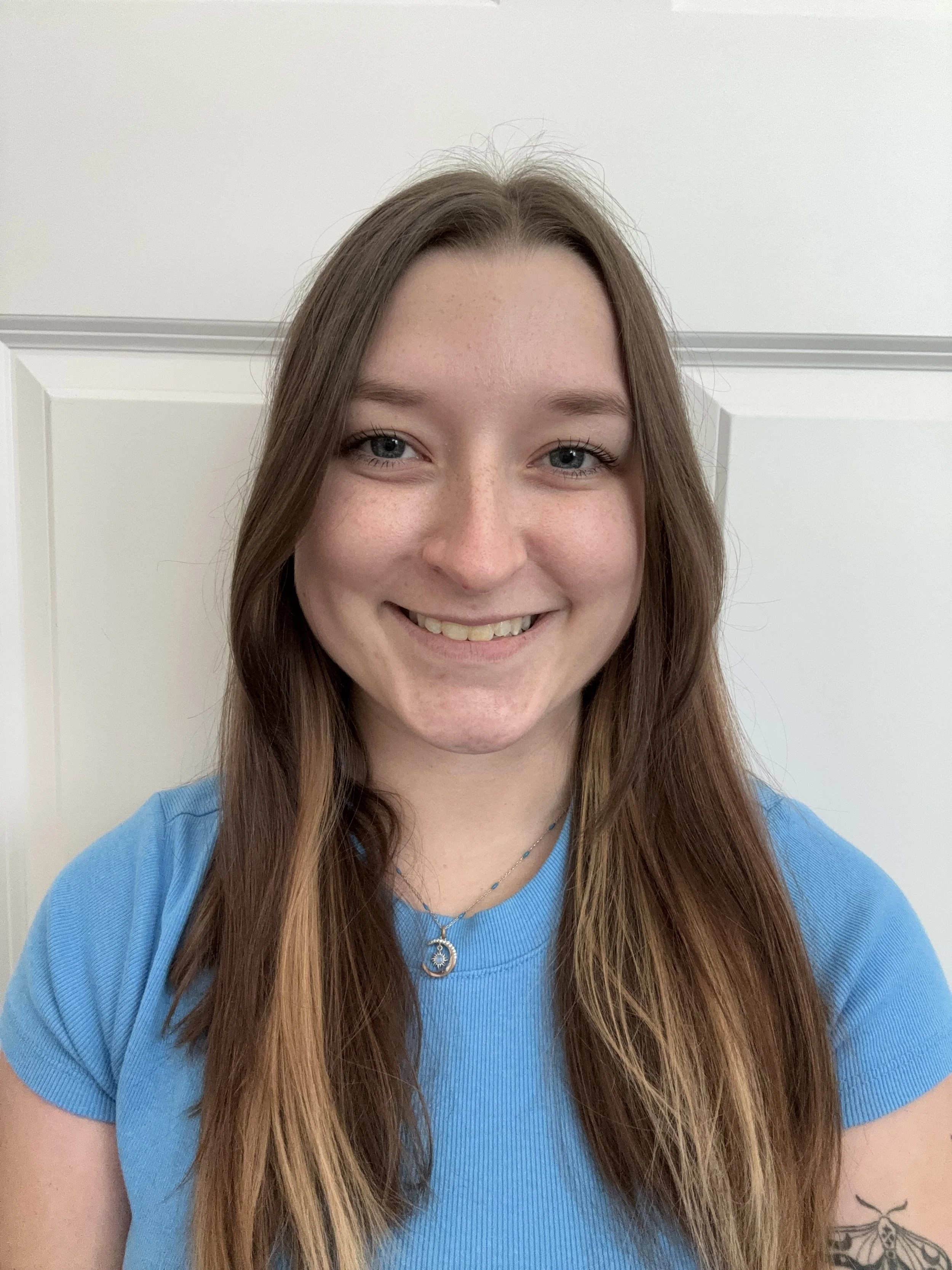 A young queer woman with long brown hair, blue eyes, and fair skin, smiling, wearing a blue T-shirt and layered necklaces, standing in front of a white door.