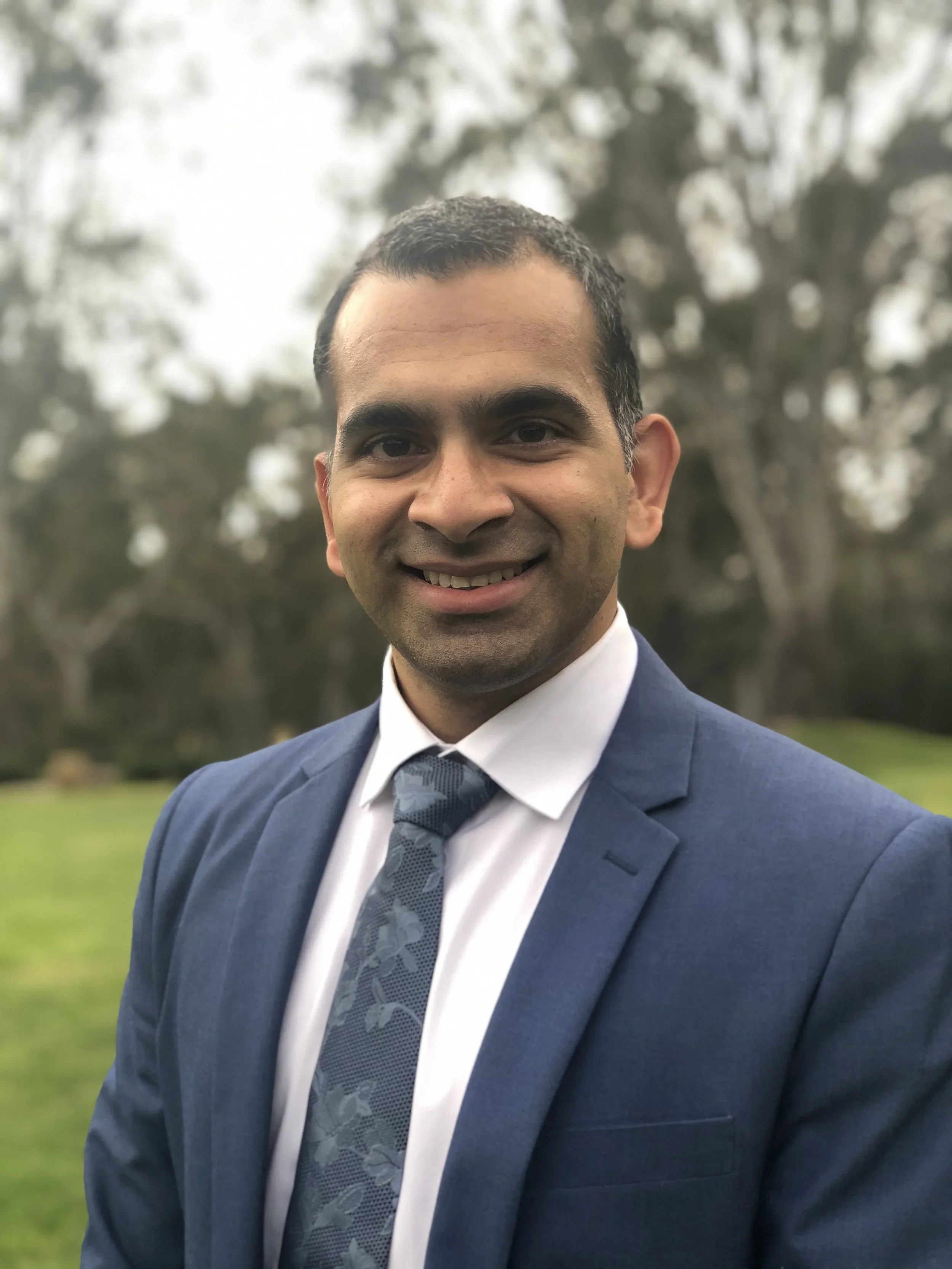 Colorectal Surgeon Dr Vignesh Narasimhan in a blue suit and patterned tie smiling outdoors with blurred trees in the background.
