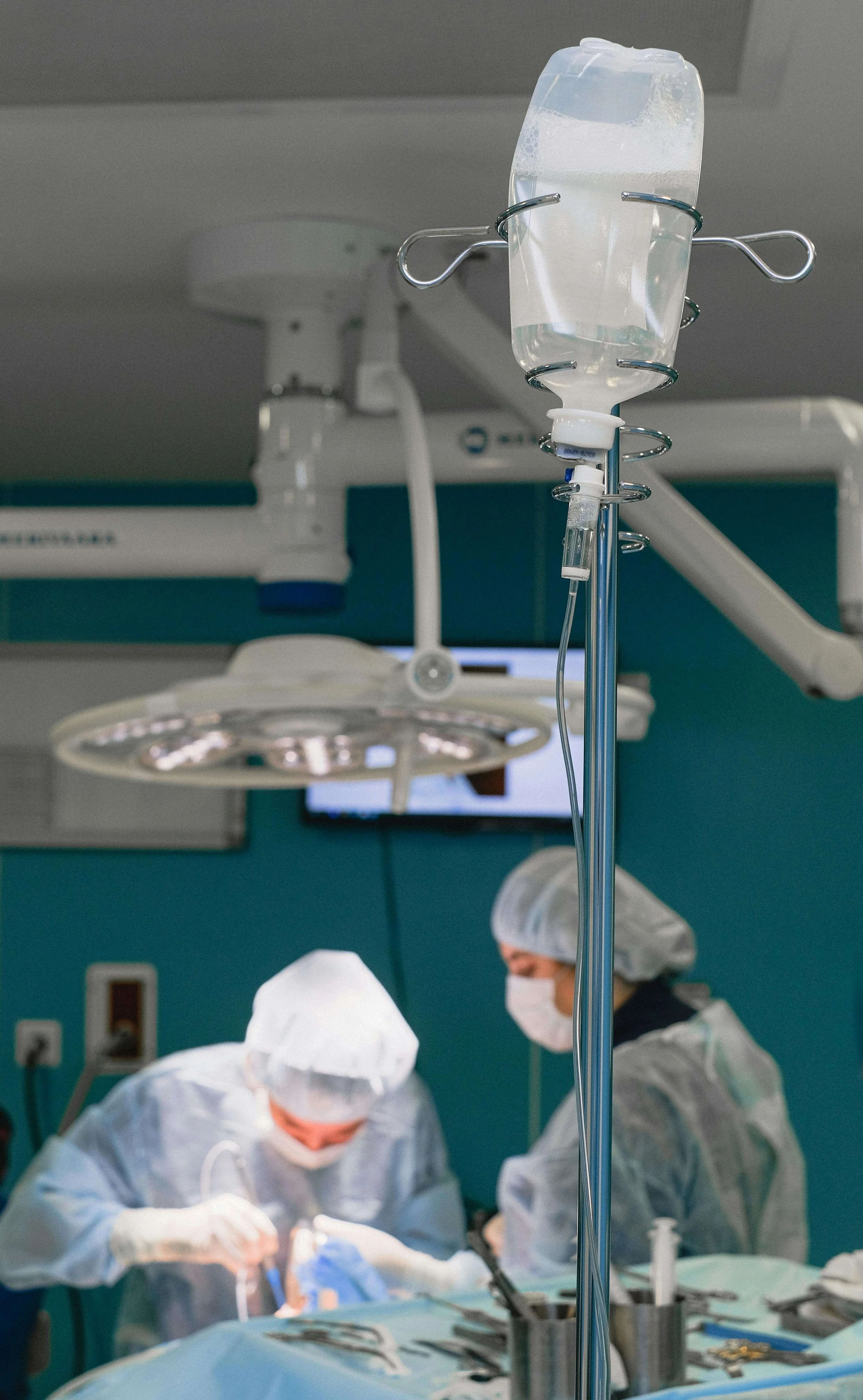Close-up of an IV drip in a hospital operating room with surgeons performing surgery in the background.