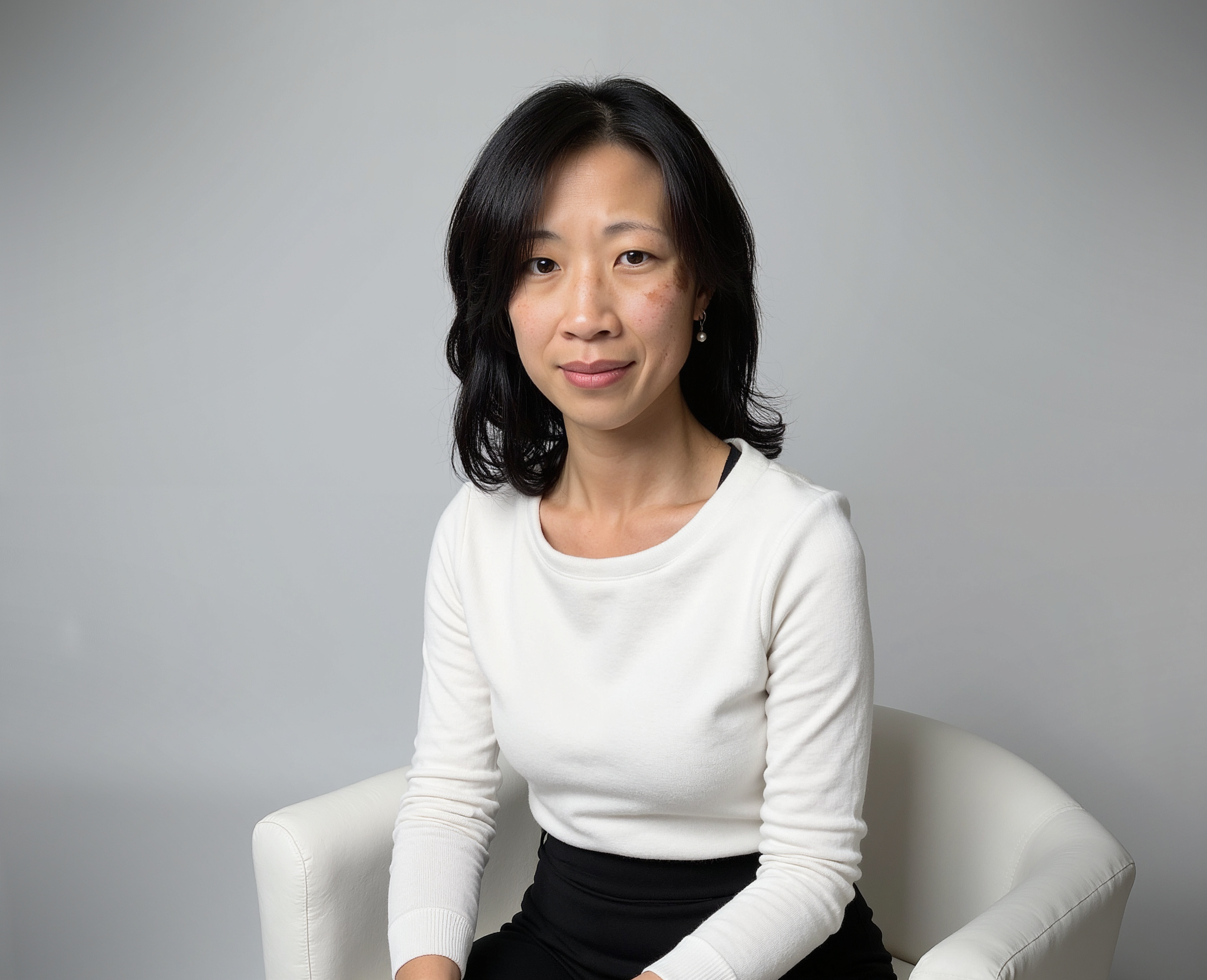 Dr Geraldine Ooi, a female surgeon with black hair and pearl earrings sitting on a white chair against a plain gray background, wearing a white long-sleeve top and black skirt.