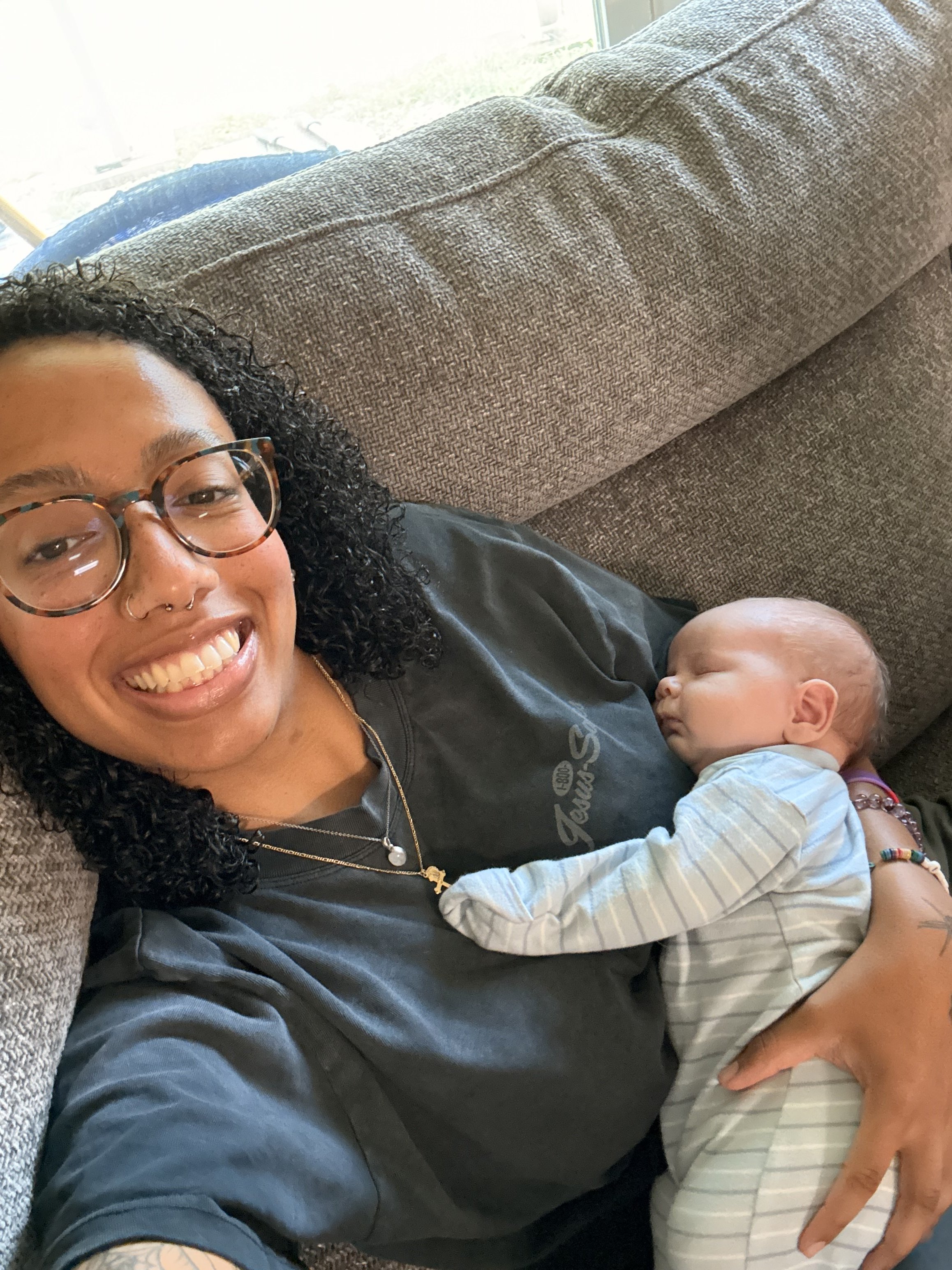 A woman with curly hair, glasses, and a nose piercing smiling while lying on a couch. She is holding a sleeping baby wearing striped pajamas.