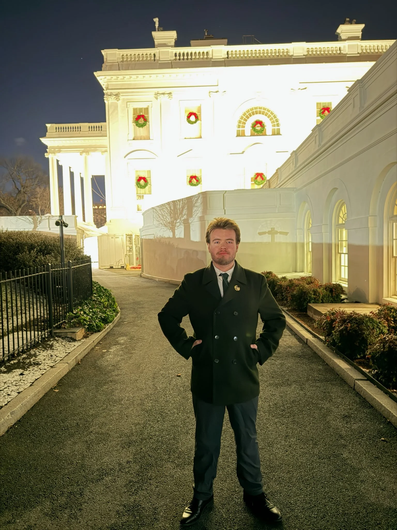 A man in a black coat and black pants standing on a dark paved pathway in front of a lit-up white building decorated with Christmas wreaths.