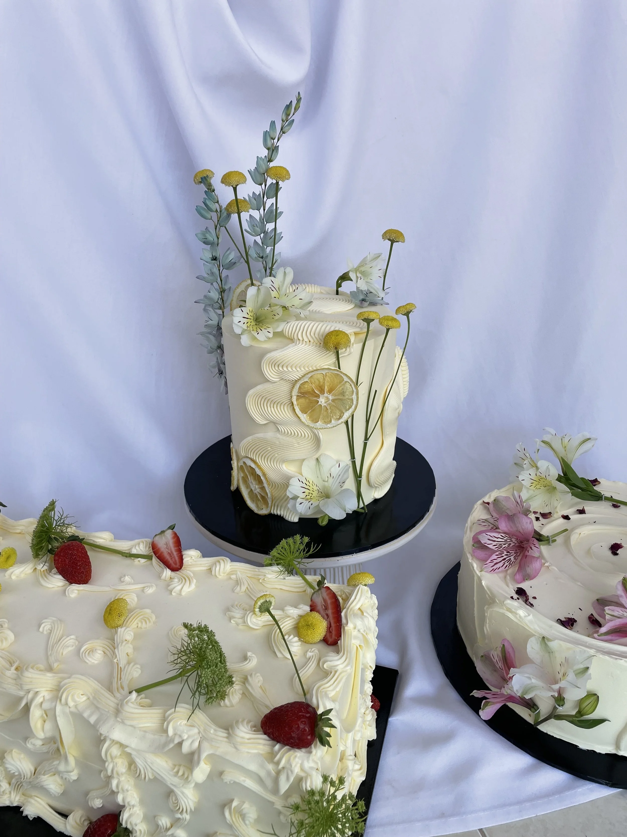 Three decorated cakes featuring fresh fruits and flowers. The tallest cake with lemon slices, white flowers, and greenery. A round cake with strawberries, yellow berries, and green garnish. A third cake with pink and white flowers.