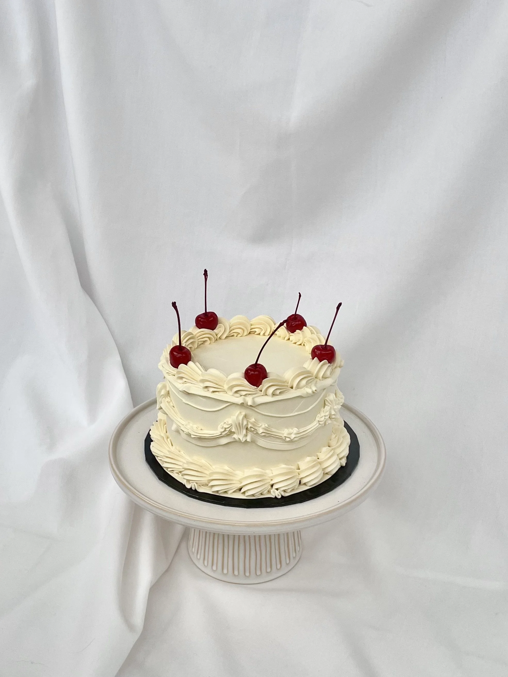 A round white cake decorated with white frosting and topped with five cherries on a white cake stand against a white background.