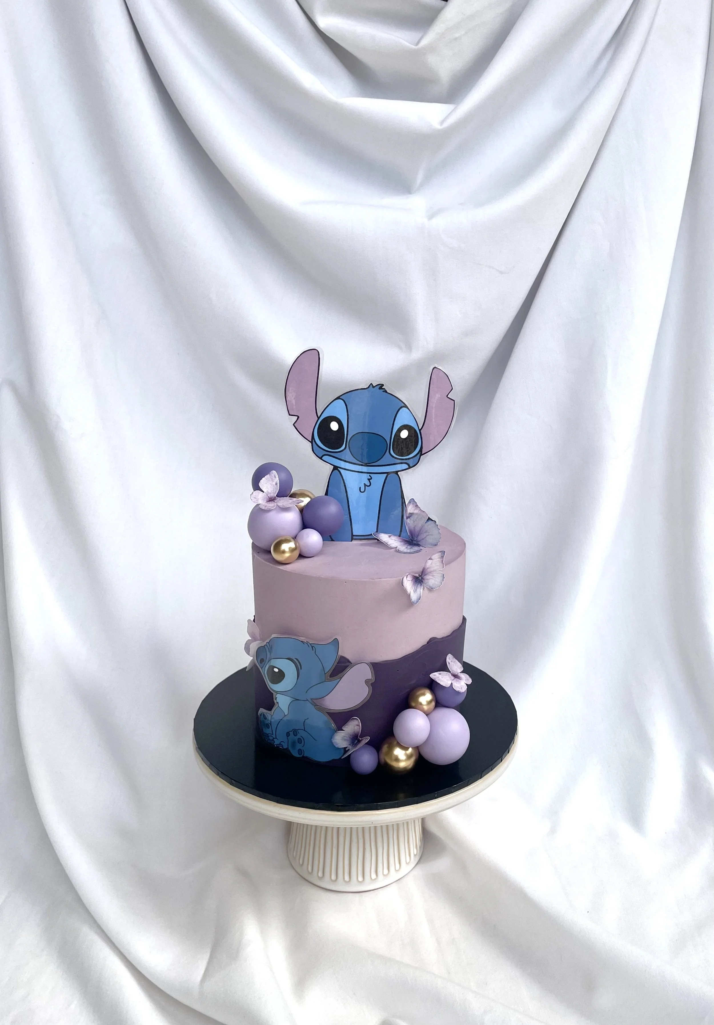 A two-tiered cake decorated with Disney's Stitch characters, purple and gold spherical accents, and purple cupcakes, placed on a black cake stand against a white fabric backdrop.