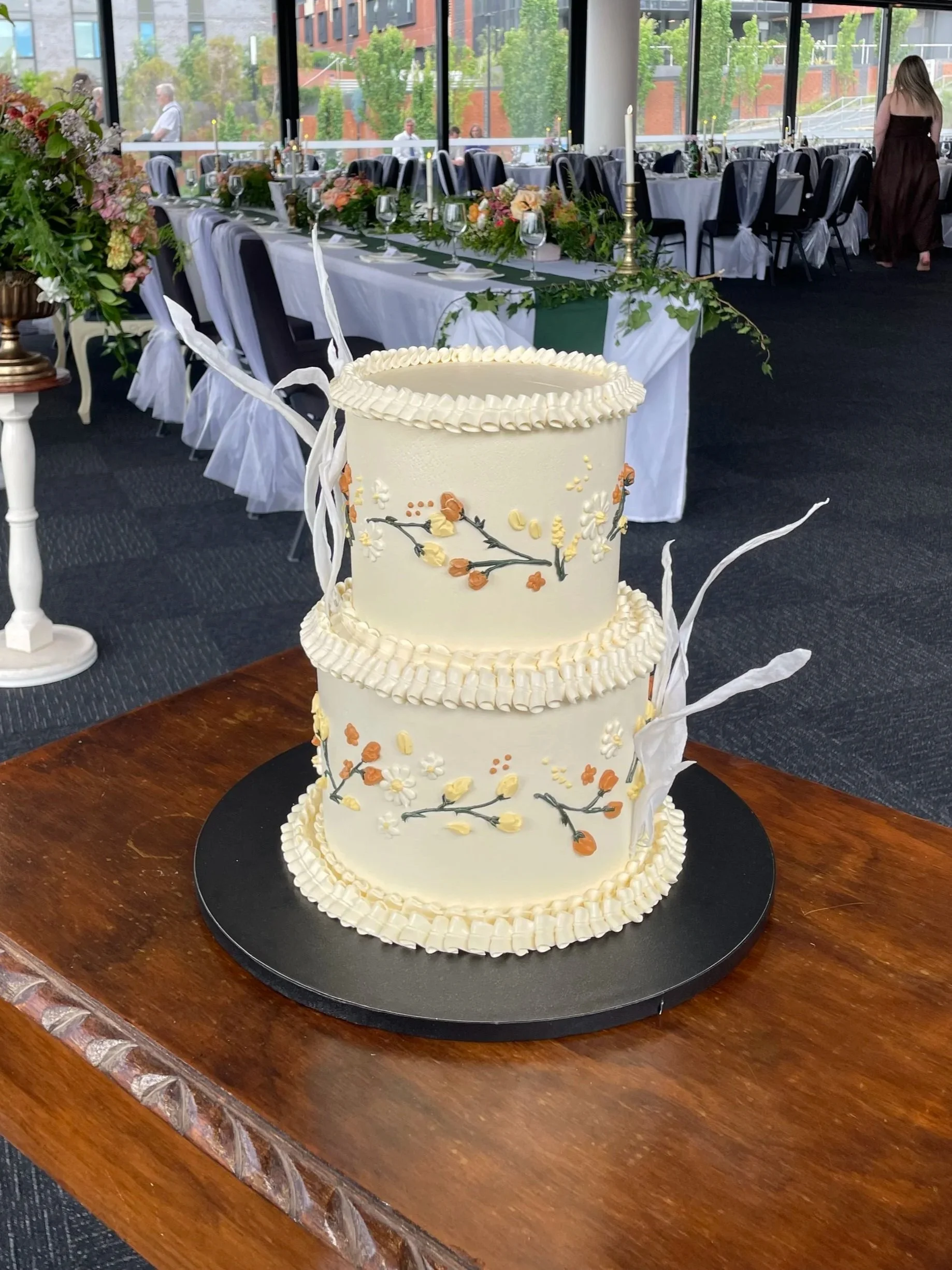 A two-tiered white wedding cake decorated with small orange, cream, and white flowers and green vines, with white chocolate accents and white ribbons, placed on a black round base on a wooden table, in a decorated banquet hall with long tables, chair
