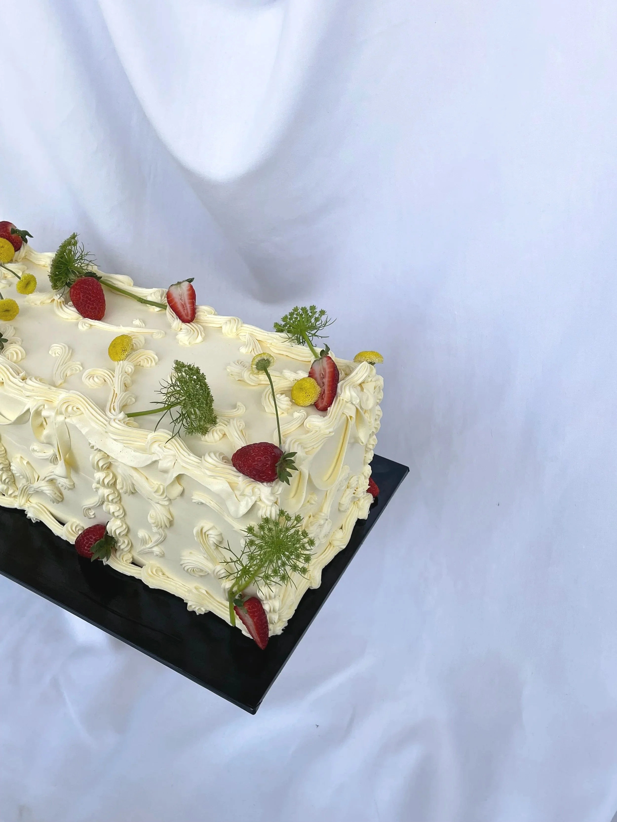 A rectangular white frosted cake decorated with strawberries, yellow billy balls, sprigs of herbs, and piped icing with decorative swirls, sitting on a black base.