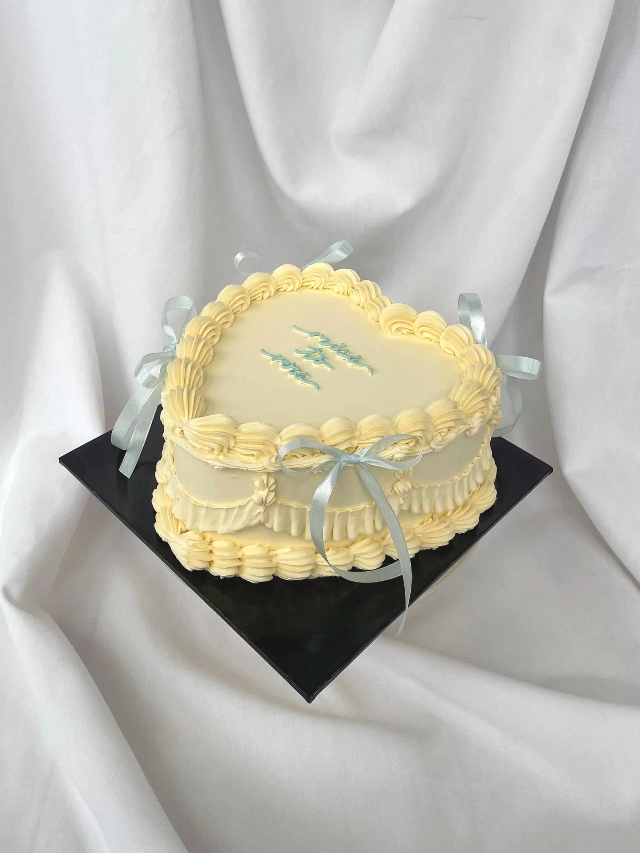 Heart-shaped cake with white and cream frosting, silver ribbons, and the words "Happy 3rd" written on top, placed on a black square base.