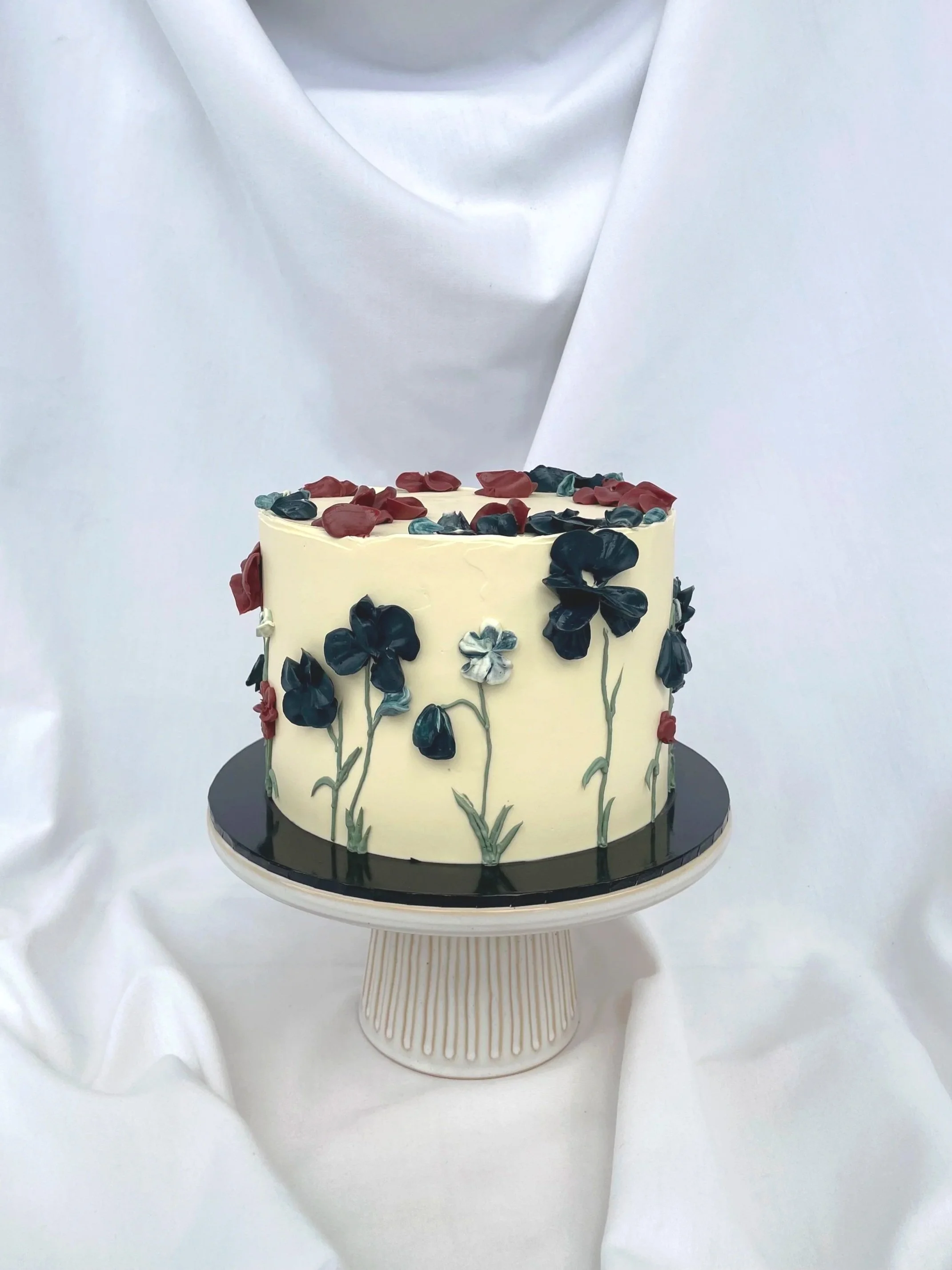 A white cake decorated with dark and light flower patterns, topped with multicolored flower-shaped decorations, on a white cake stand against a white fabric background.