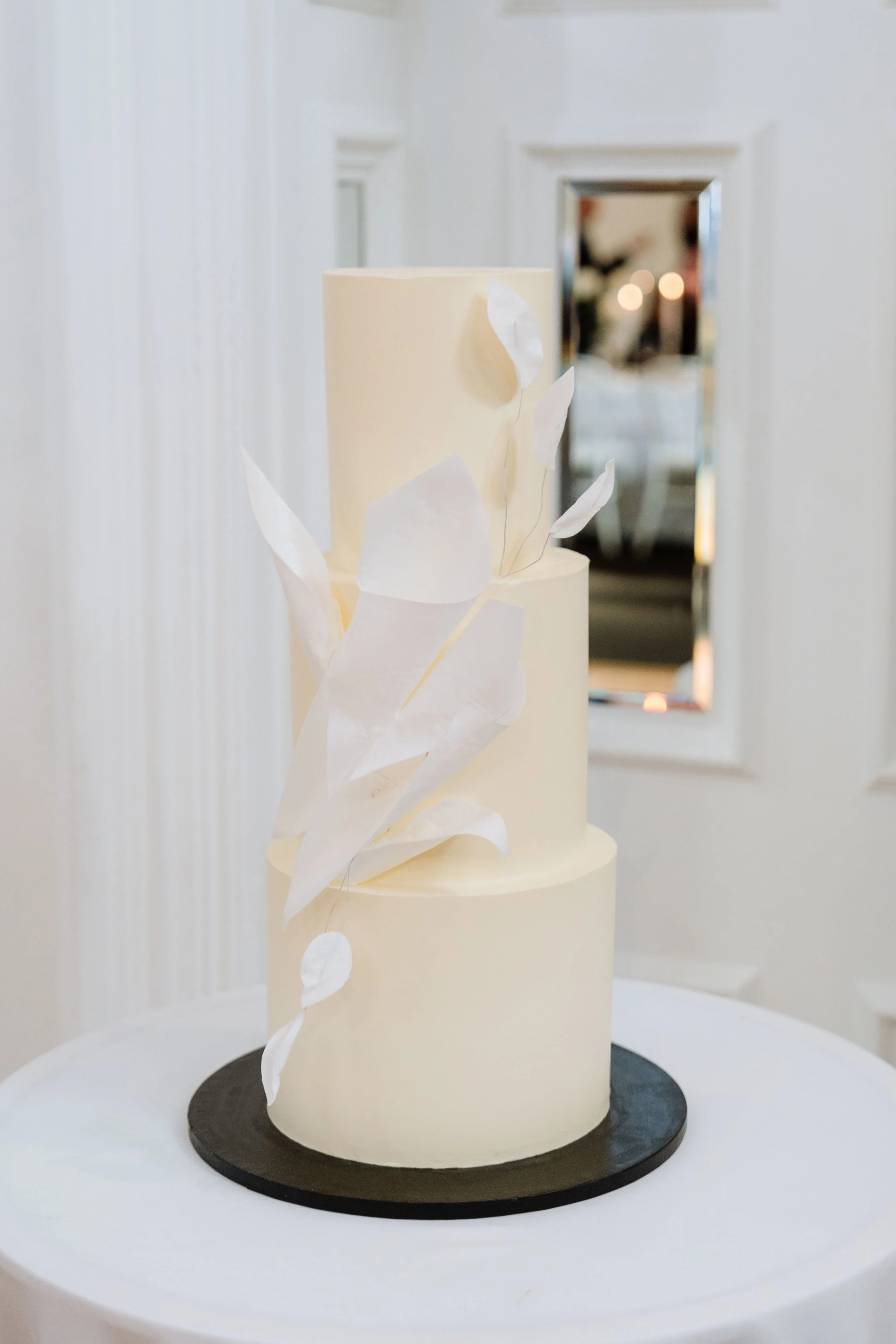 Three-tier white wedding cake with decorative white paper leaves, on a black cake board, in front of a mirror with a white wall background.