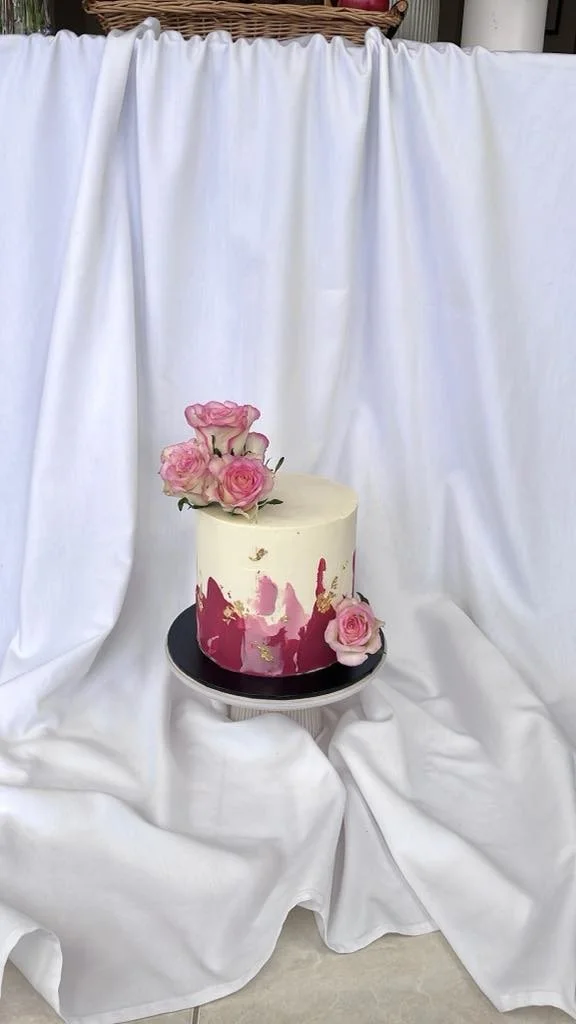 A single-tier white cake with pink and gold accents, decorated with pink roses, on a round black cake stand in front of white draped fabric.