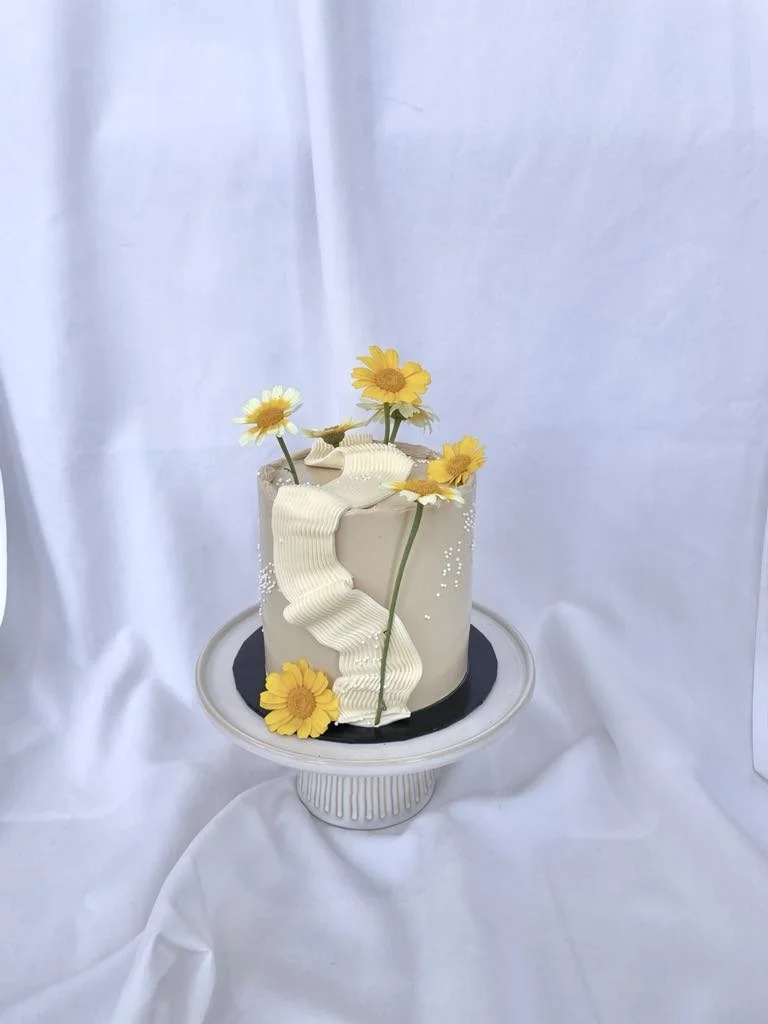 A tall, white cake decorated with yellow flowers and a white ribbon, displayed on a white cake stand against a white background.