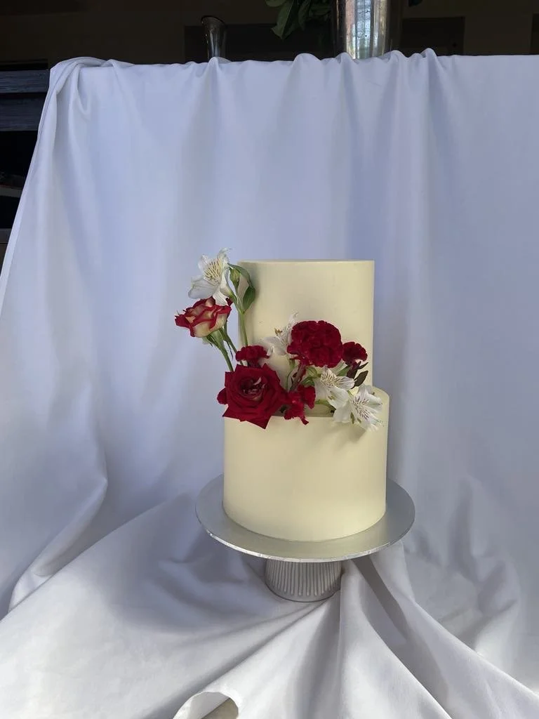 Two-tiered cream-colored wedding cake decorated with red and white flowers, placed on a round silver cake stand with white drapery and green plant in the background.
