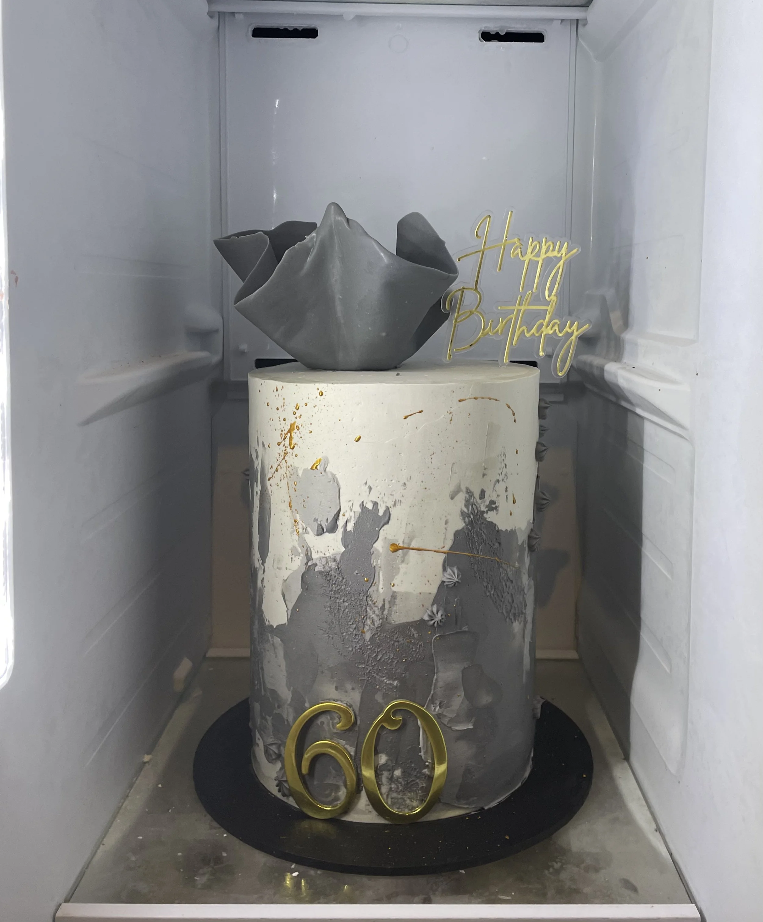 A birthday cake decorated with gray and gold accents, displaying the number 60, with a black base inside a refrigerator. The cake has a gray, abstract flower topper and a gold 'Happy Birthday' sign.