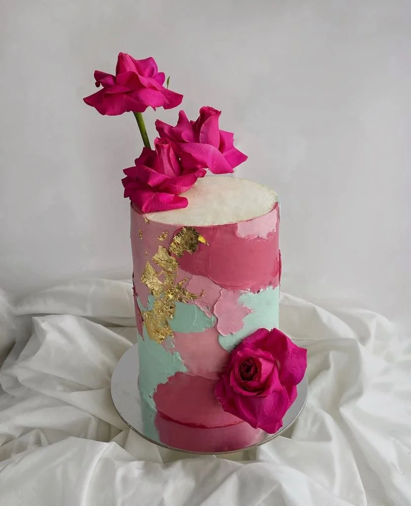 A tall, cylindrical pink, blue, and white marble-patterned cake decorated with pink roses and gold accents, placed on a round white base with white fabric underneath.