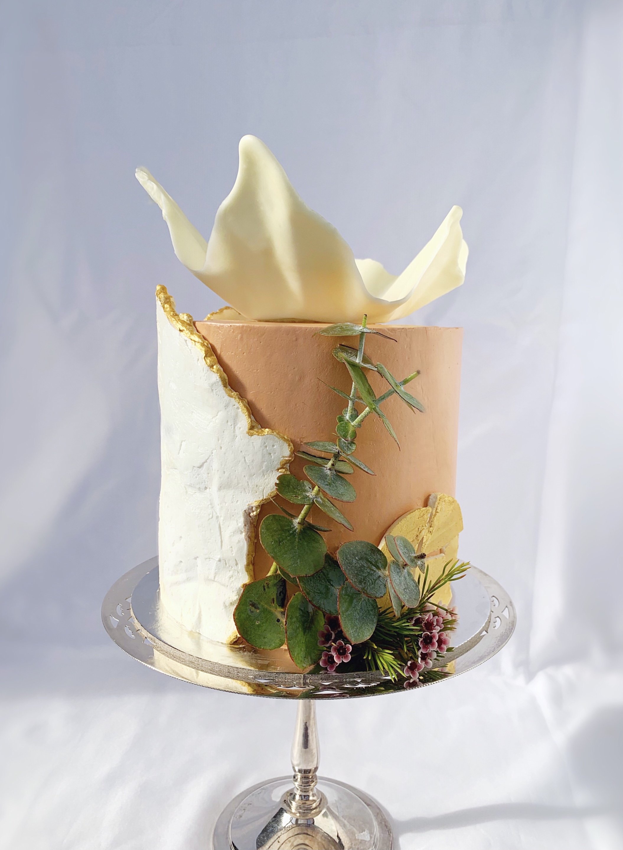 Elegant cake with a light pink and white exterior, decorated with greenery, a white chocolate sculpture on top, and gold accents, displayed on a glass cake stand.
