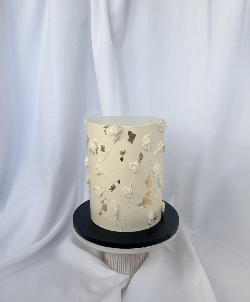 A tall cylindrical white cake decorated with small white and gold floral and butterfly accents, placed on a black cake board on a round stand, with white fabric draped in the background.
