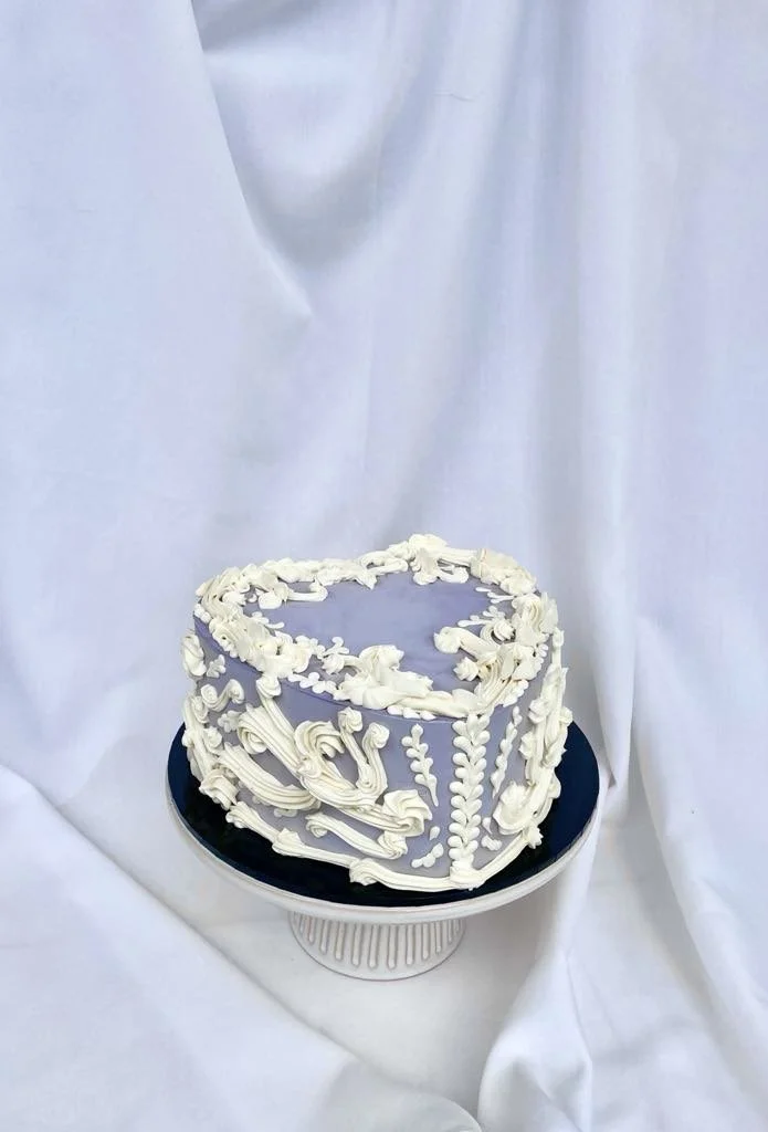A heart-shaped cake decorated with white ornate piping and designs on a black cake board, with a white backdrop.
