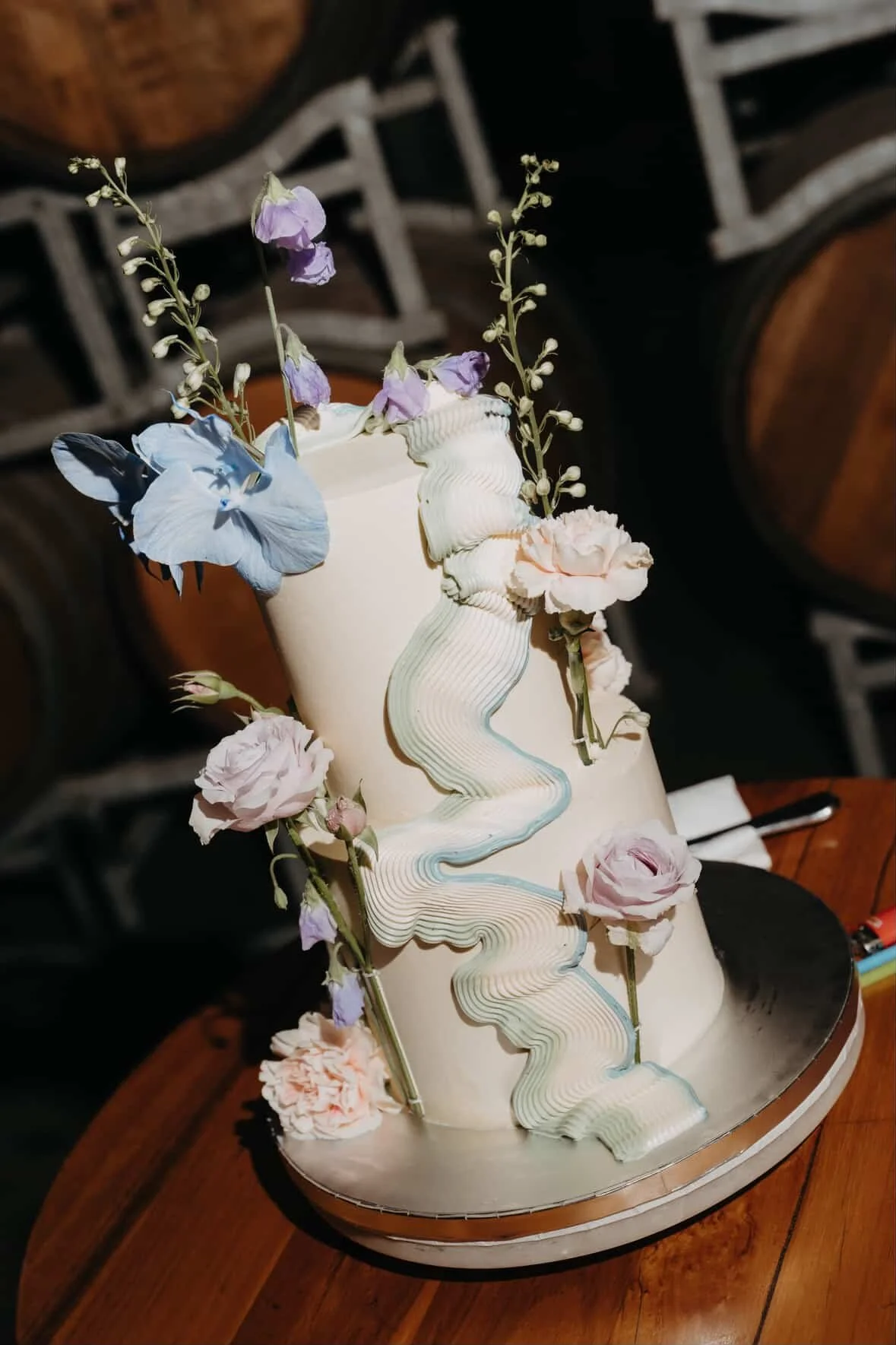 A tall, white, cylindrical birthday cake decorated with pastel-colored flowers, including pink and purple blooms, and a long, striped, edible cascade draping down the side.