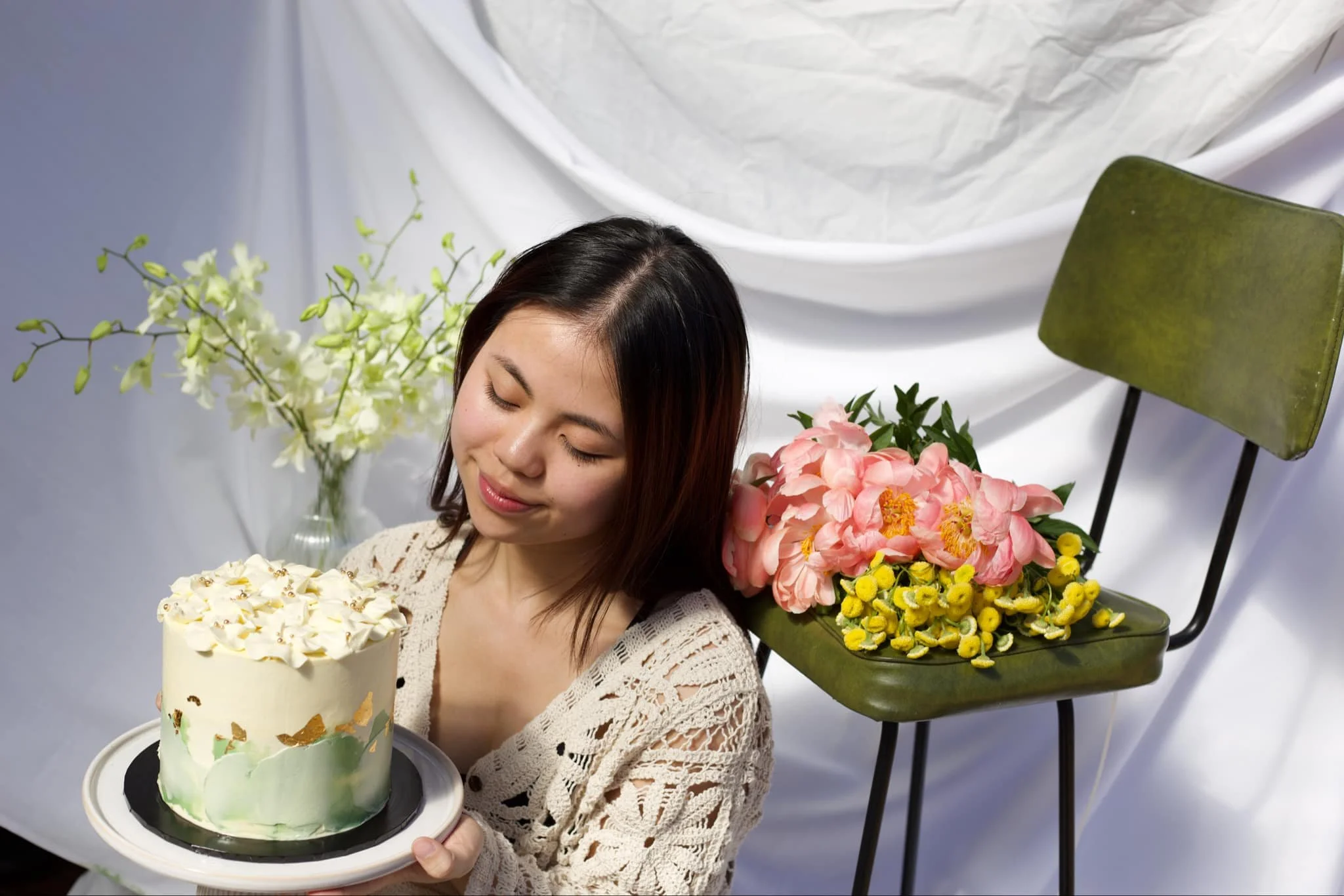A woman with shoulder-length dark hair holding a cake with white frosting and cream decorations. She is sitting near a green chair with pink and yellow flowers placed on it, against a backdrop of white fabric and flower arrangements.